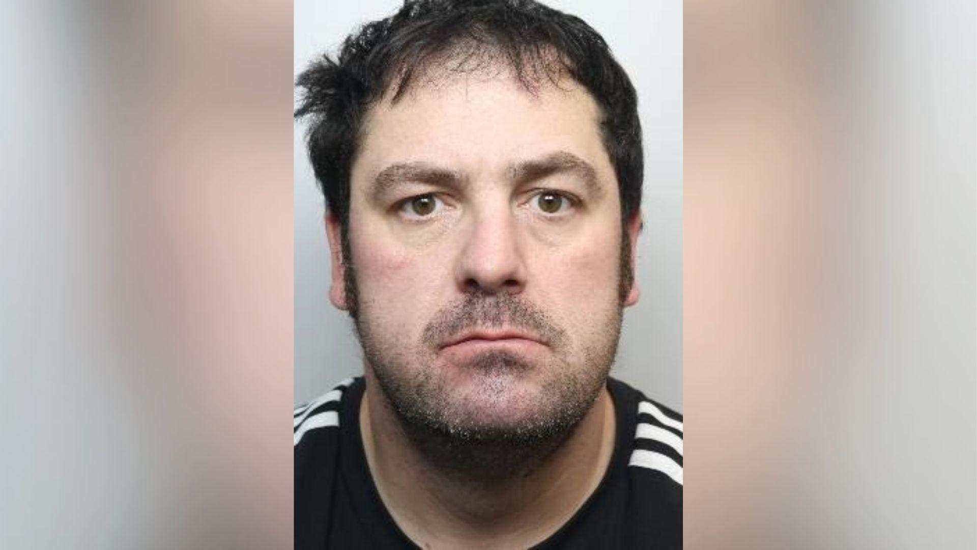 A police mugshot of Trevor Lockwood. He has an oval face, a thin mouth which is slightly downturned, black hair and sideburns, and dark stubble. He is wearing a black T-shirt with white Adidas stripes on the shoulders.