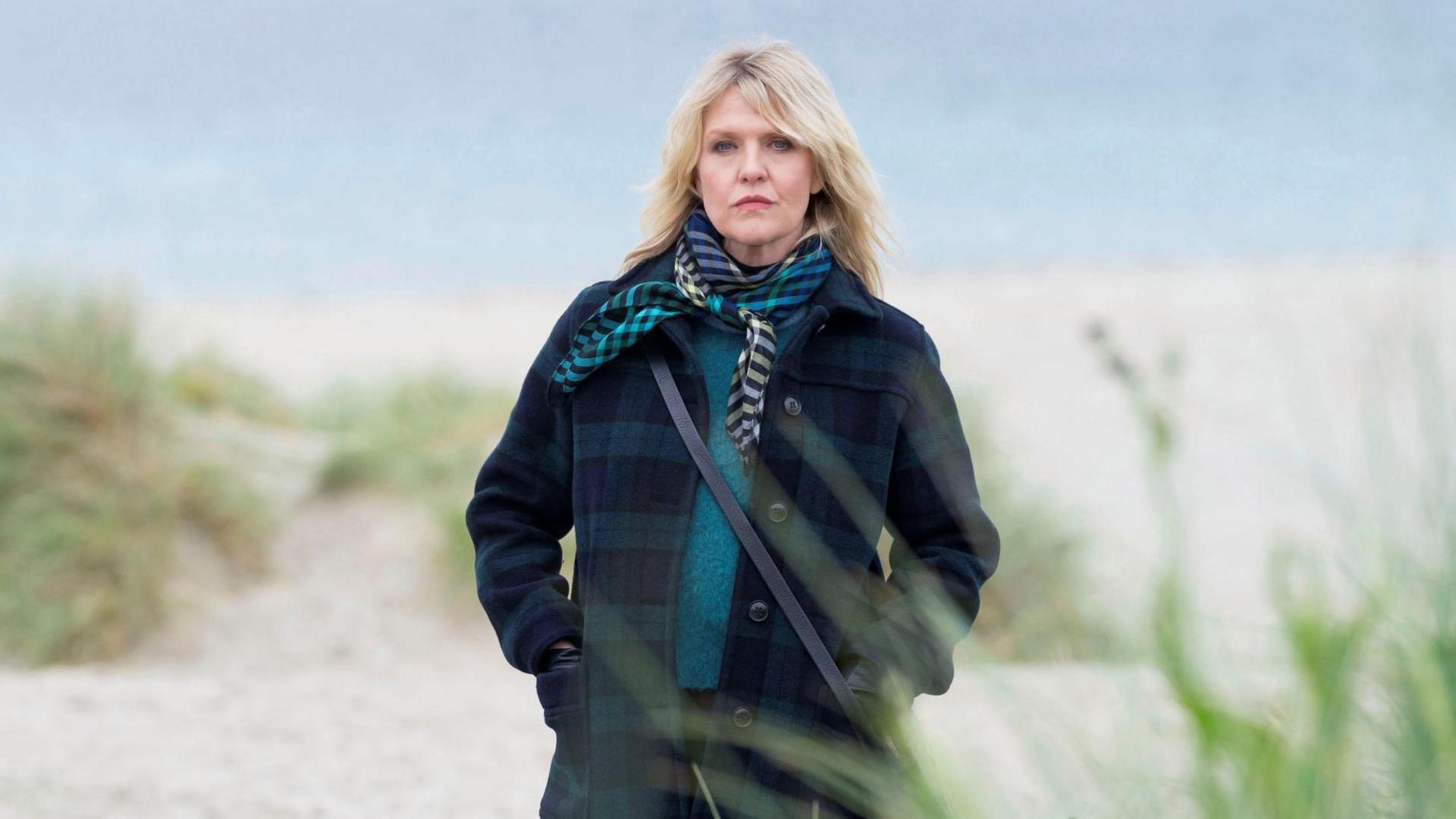 Ashley Jensen stands on a deserted Shetland beach, the sea grey behind her. She is wrapped up in a thick green and navy tartan wool coat with a teal woolly jumper and a patterned scarf. Her hands are in her pockets as she looks moodily at the camera.