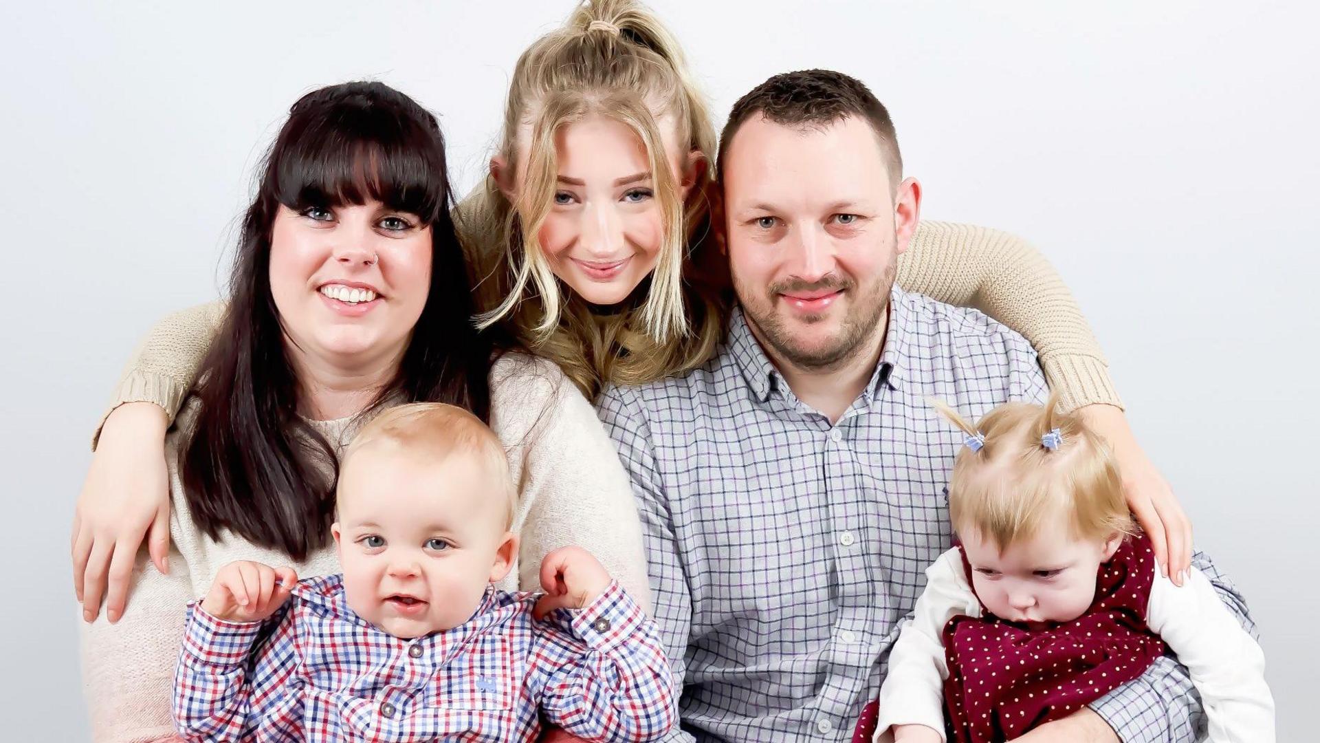 A family of five smile at the camera. Two babies are held in the arms of a woman and a man while an older girl leans over in between them with her arms around the man and woman. They are all smiling at the camera.