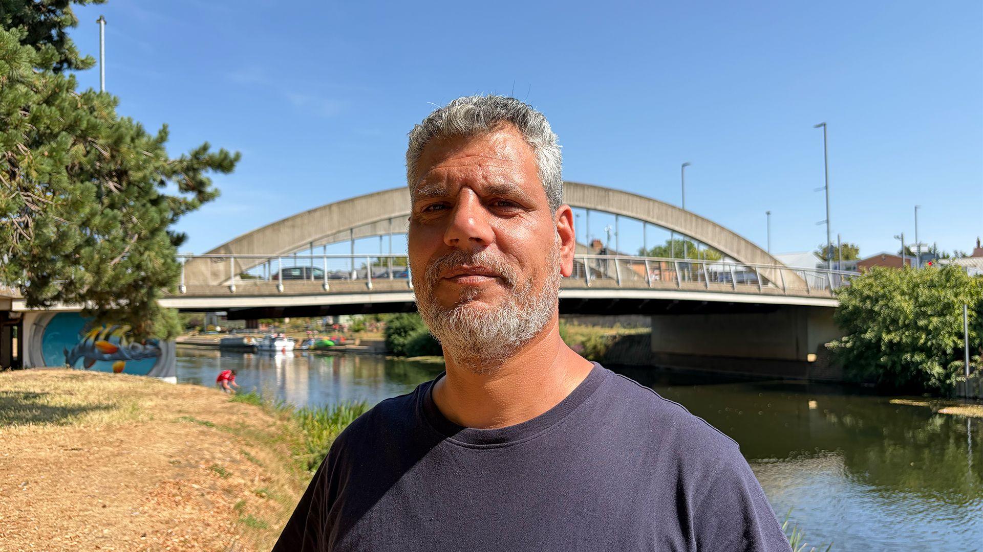 Samer Jouha standing by the River Avon in Evesham, close to where he pulled the woman from the water. It is a sunny day and behind him is a bridge. There are also trees behind him on both banks of the river.
 