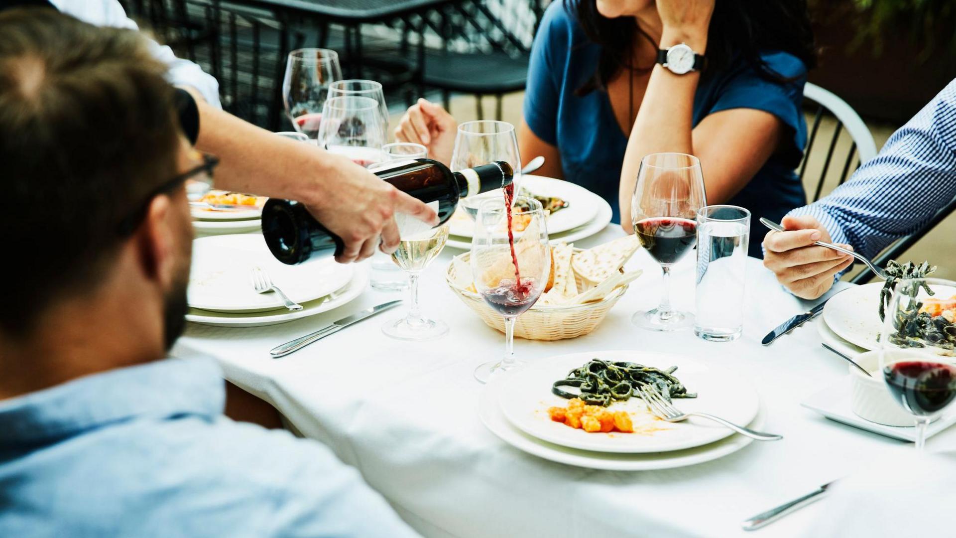 Dining table with red wine being served. There is a white linen table cloth and finished plates of food on white plates.