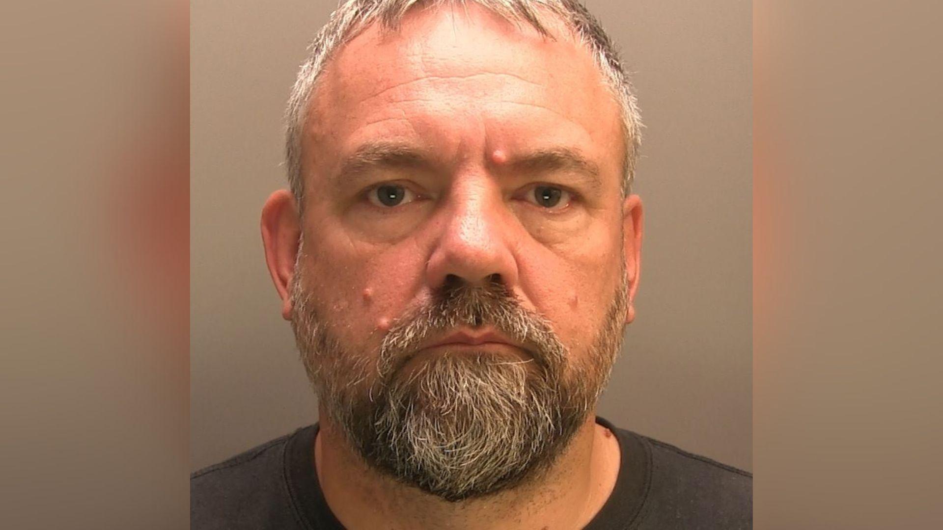 Brett Peterson has short grey hair, a beard and moustache and looks into the camera lense. He is wearing a black T-shirt.