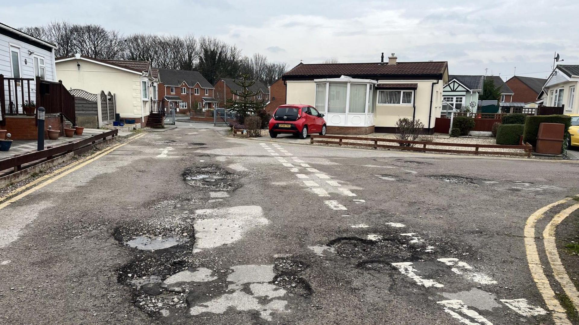 A junction of roads on a mobile home park. There are some big potholes in the foreground, and stretching away into the distance. A cream coloured mobile home with a brown roof and front porch sits on the curve of the junction with a small red car parked in front. There are double yellow lines running along the sides of the road. Other mobile homes are visible on the left-hand side and some gates stand open in the background with red brick houses visible beyond. 