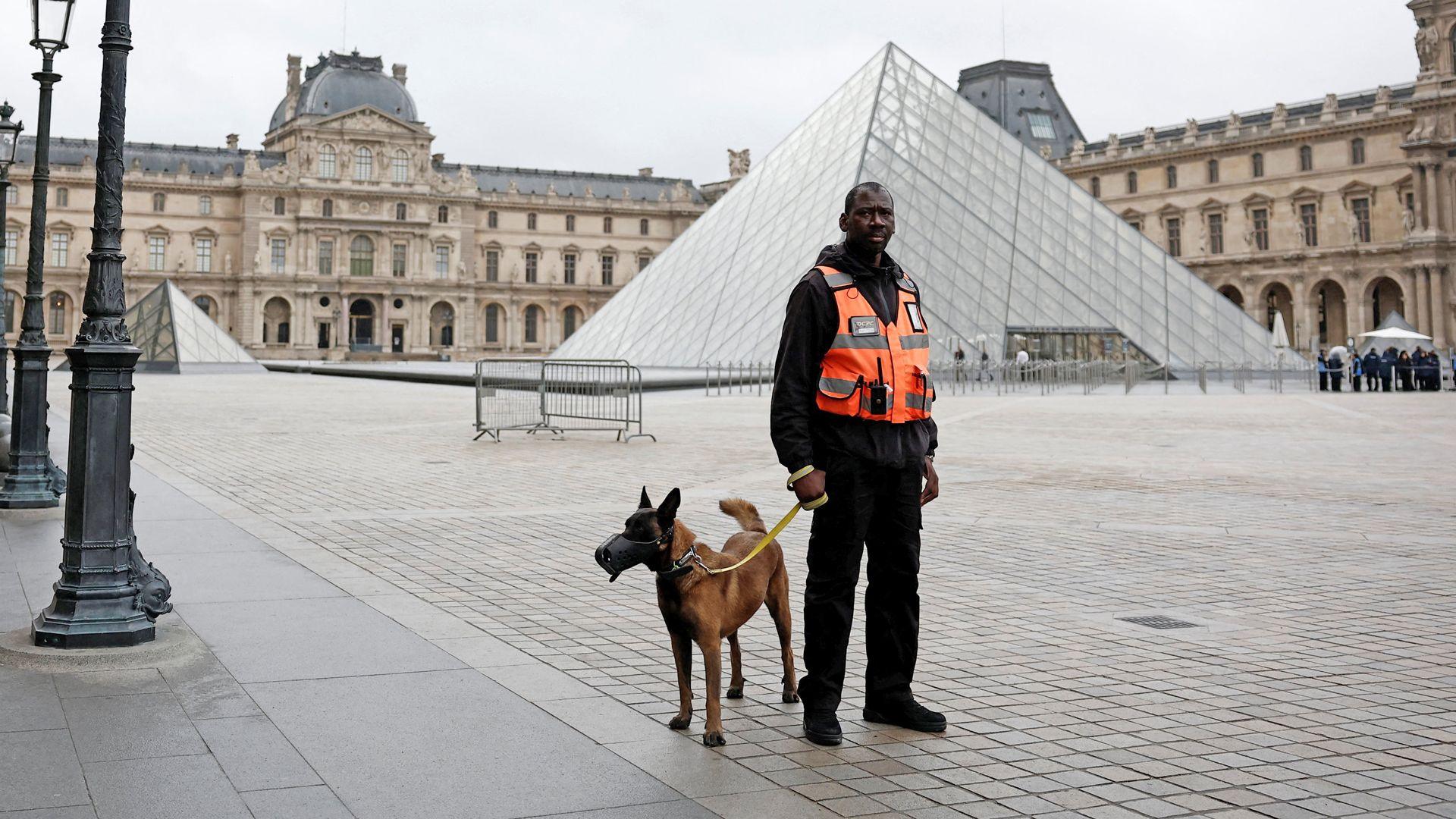 A security guard and dog stand outside the iconic 3D triangle exterior of the Louvre in Paris, which is shown looking very empty as it remains closed.