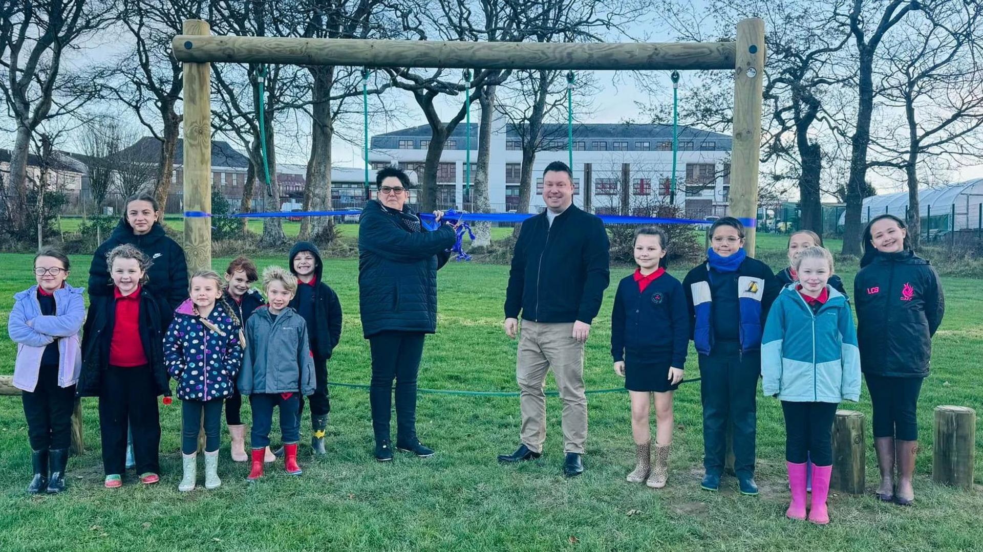 Primary school children standing in front of the wooden play area. There is a rope balancing beam with rope handles, two adults stand in the middle about to cut a blue ribbon hung between the wooden posts at either side of the balancing beam.