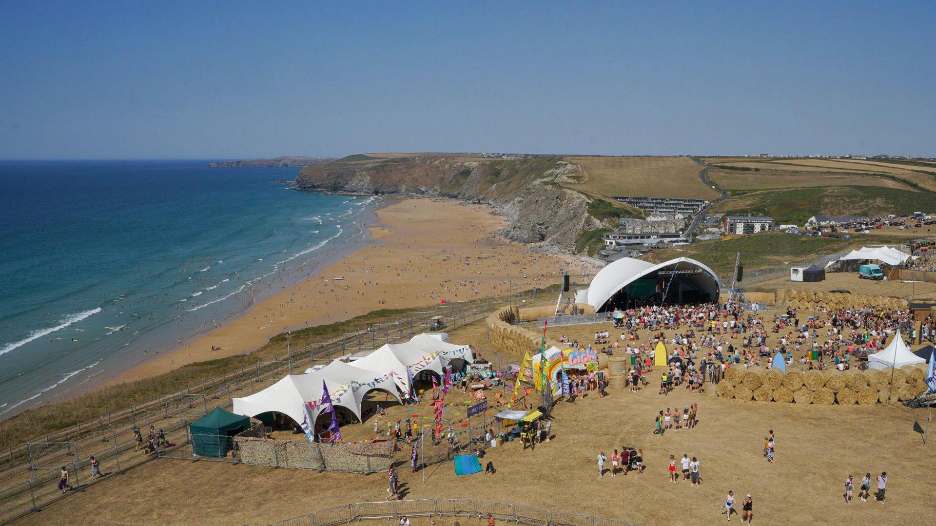 A large outdoor festival near a beach, featuring white tents, colorful structures, and crowds of people on a grassy area. A stage with a white canopy is visible in the background, while attendees enjoy the beach along a scenic coastline with cliffs and hills.