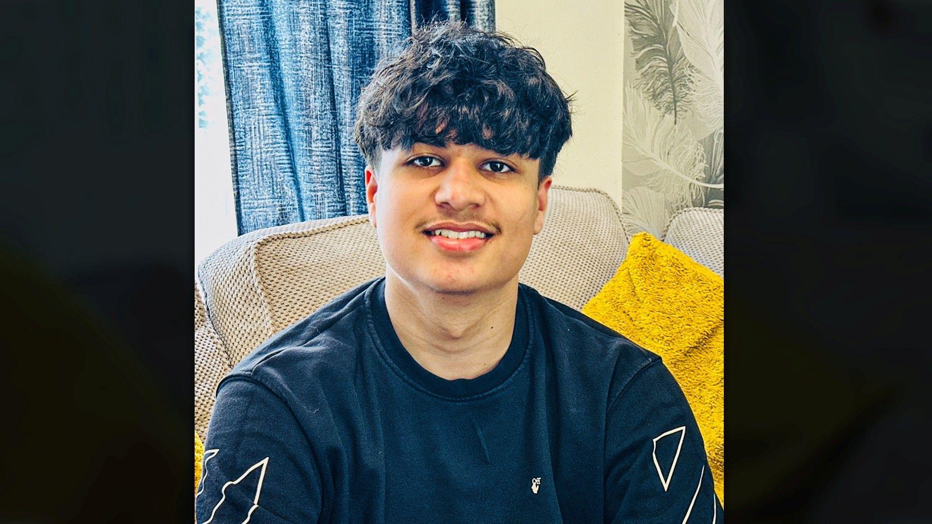 A young antheral   sat connected  a beige sofa with a yellowish  cushion beside them. He is wearing a acheronian  sweatshirt with achromatic  geometric designs, with a bluish  curtain and leaf‑patterned wallpaper down  them.