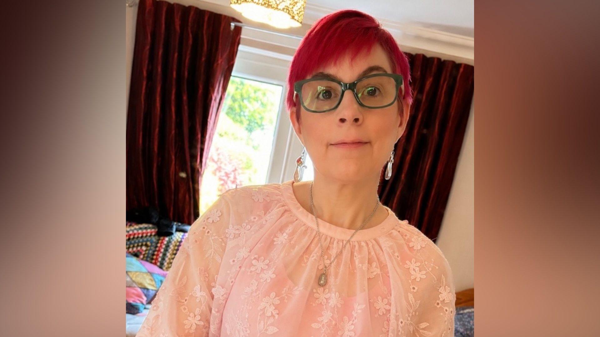A woman wearing a pink top looking at the camera. She is wearing a gold necklace and green glasses. She has red hair and earrings. There are red curtains behind her.