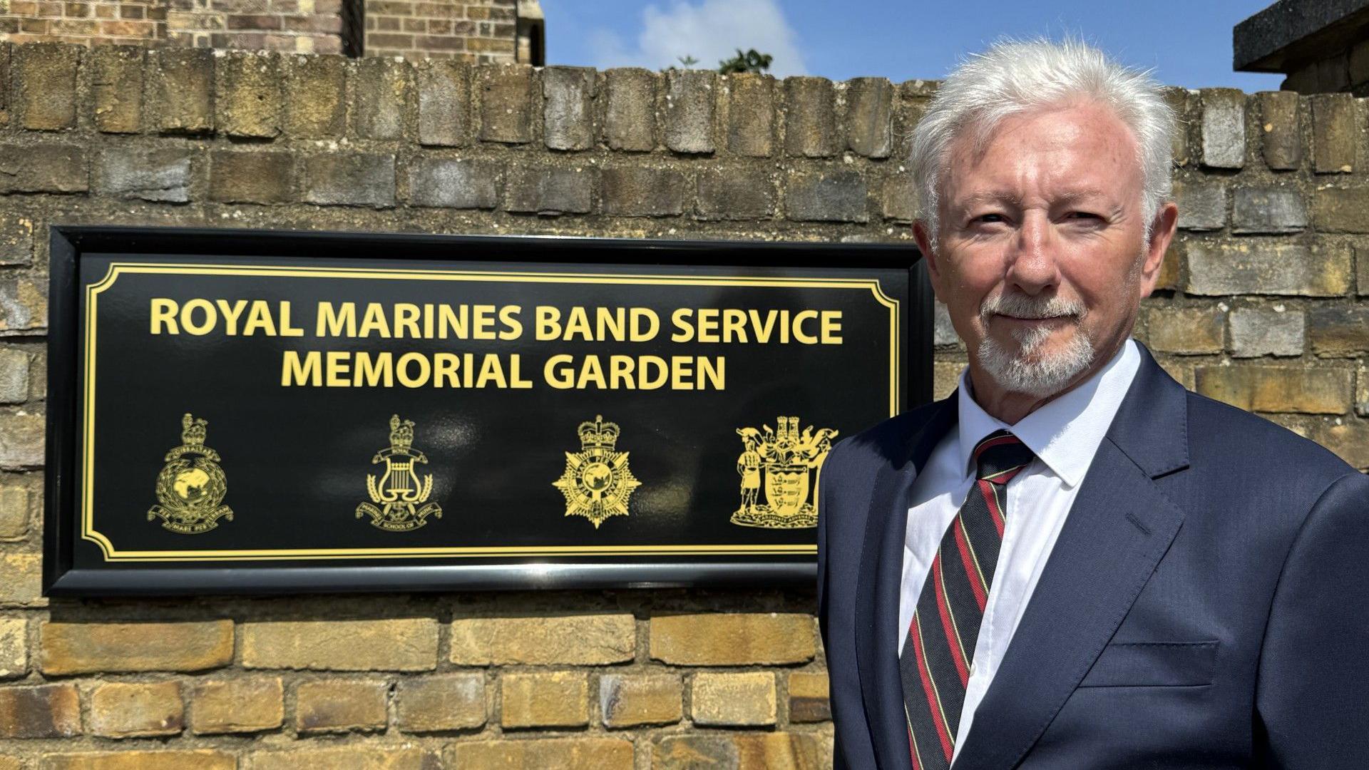 A man wearing a suit stood in front of a brick wall with a black sign with the words ROYAL MARINES BAND SERVICE MEMORIAL GARDEN in gold letters