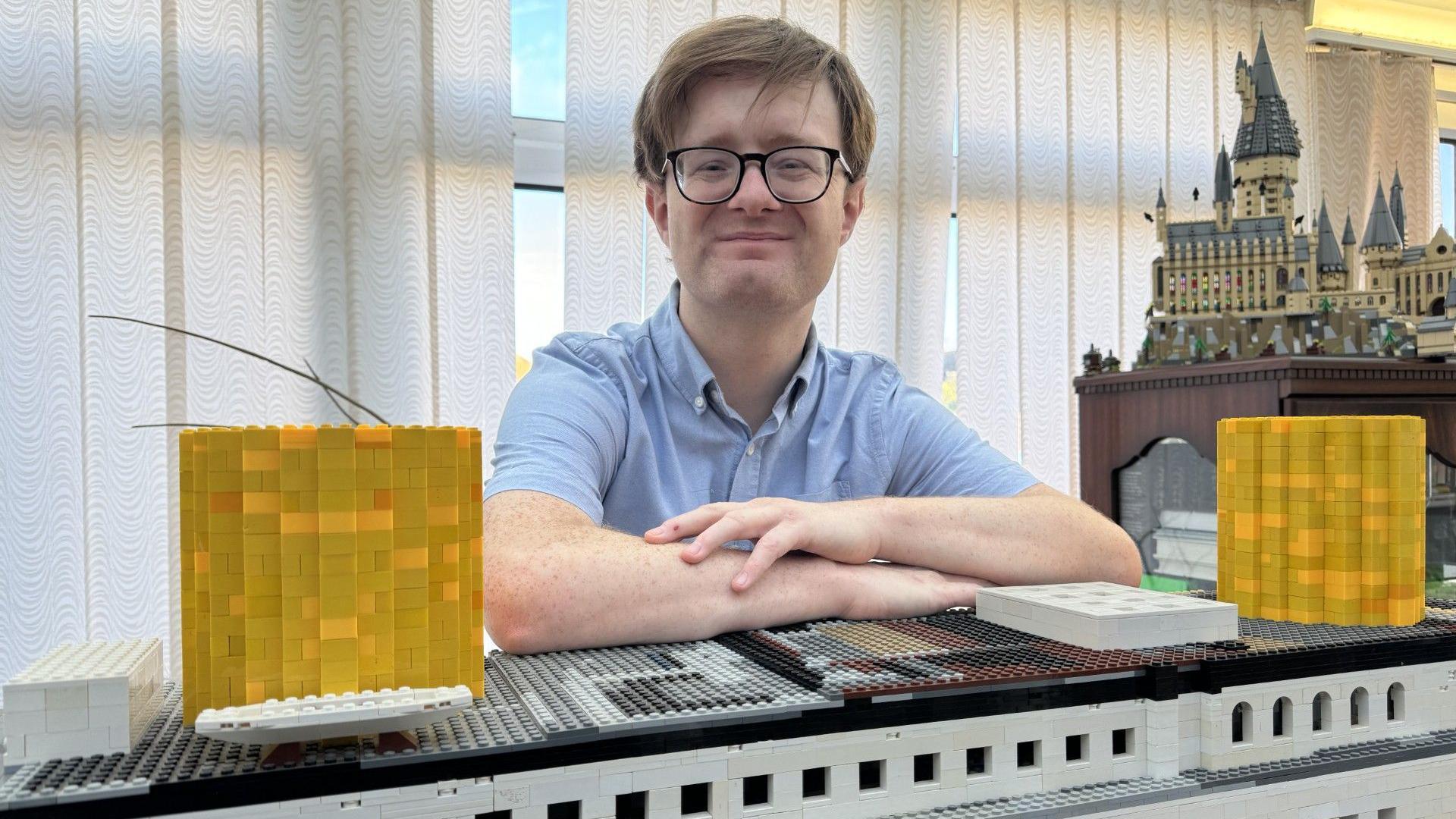 Jack Moore (a man with glasses and his hair in a curtain style, wearing a blue shirt) looks directly into the camera and leans on his Lego creation of the titanic