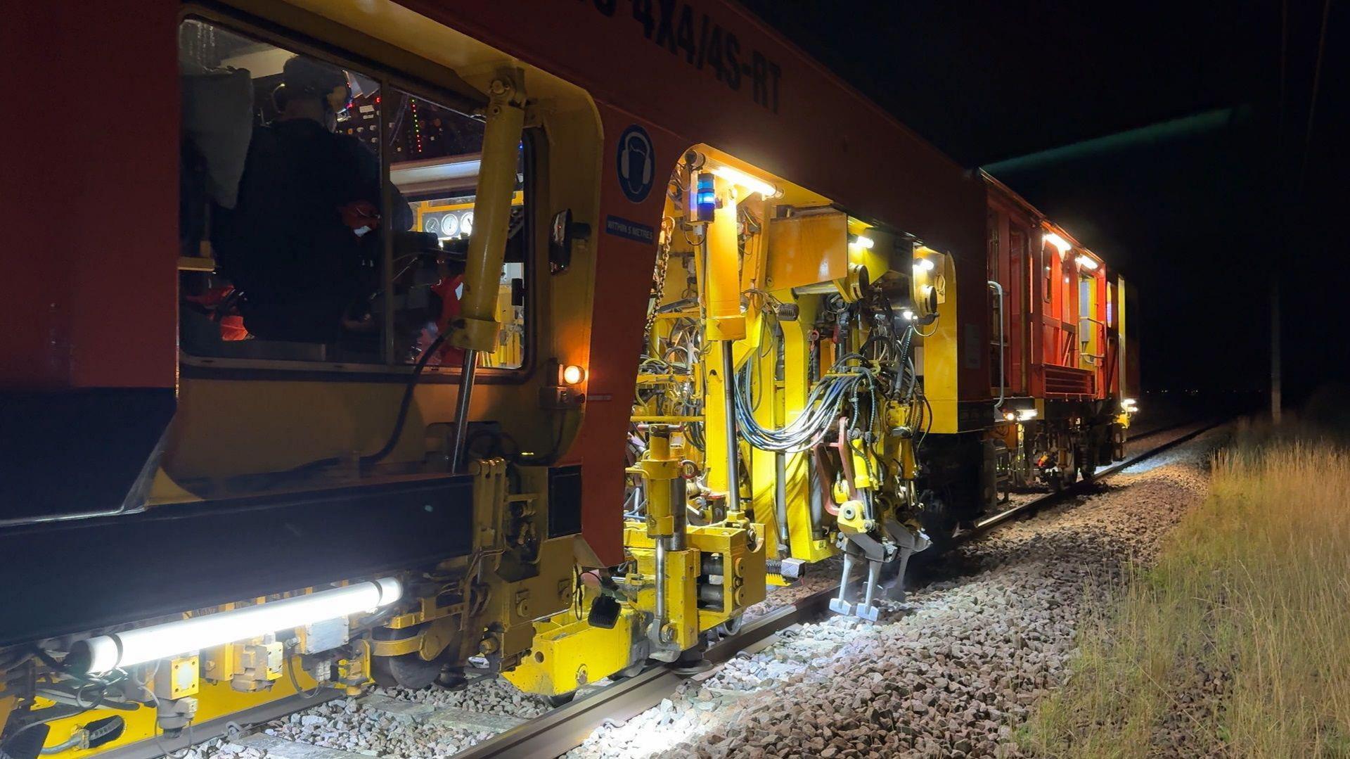 A tamper machine being operated at night on a railway track. You can see large stones near the track being pressed by a robotic-type arm