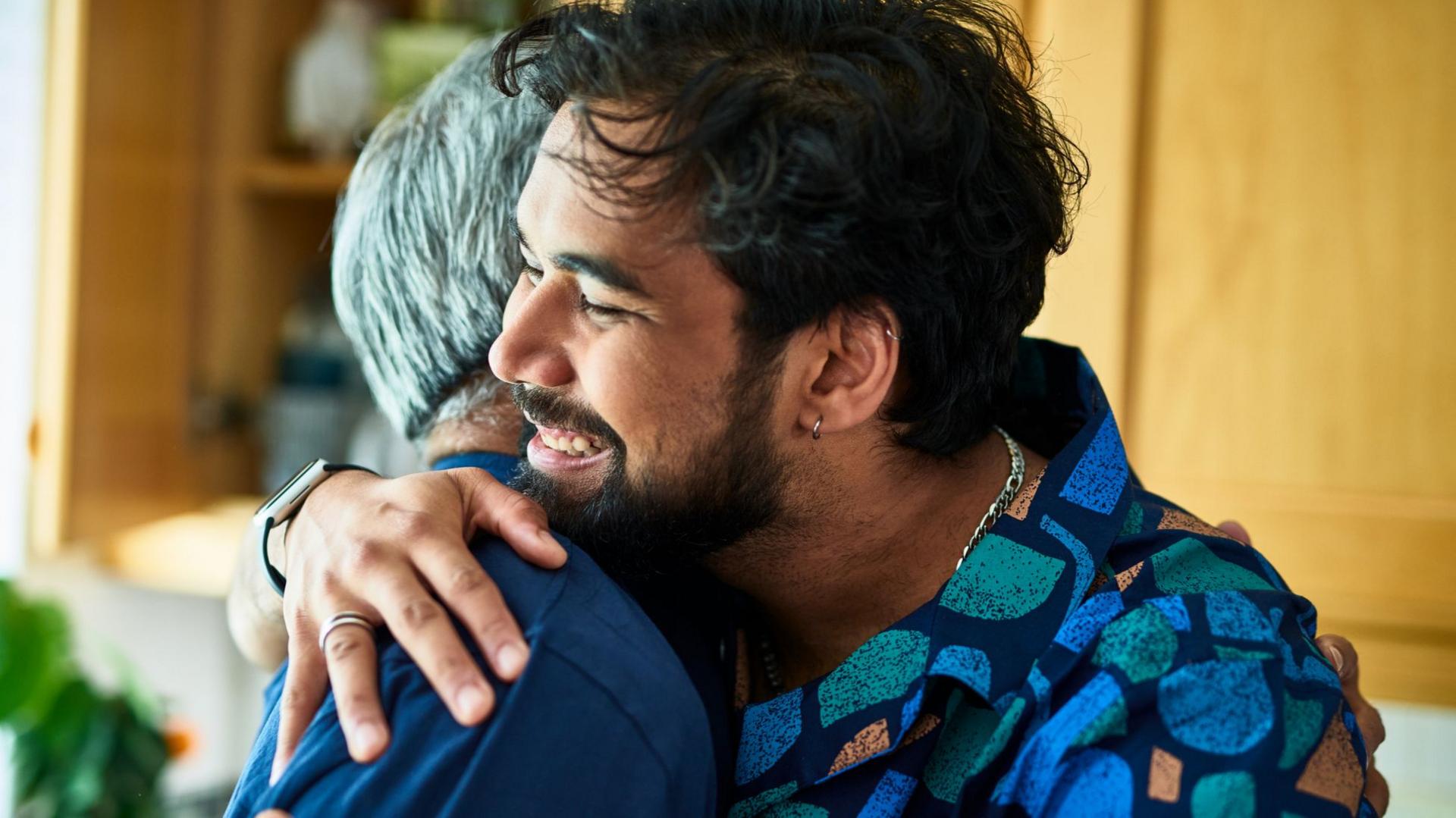 A man with dark hair is hugging somebody else with grey hair whose face we cannot see. The man we can see is smiling.