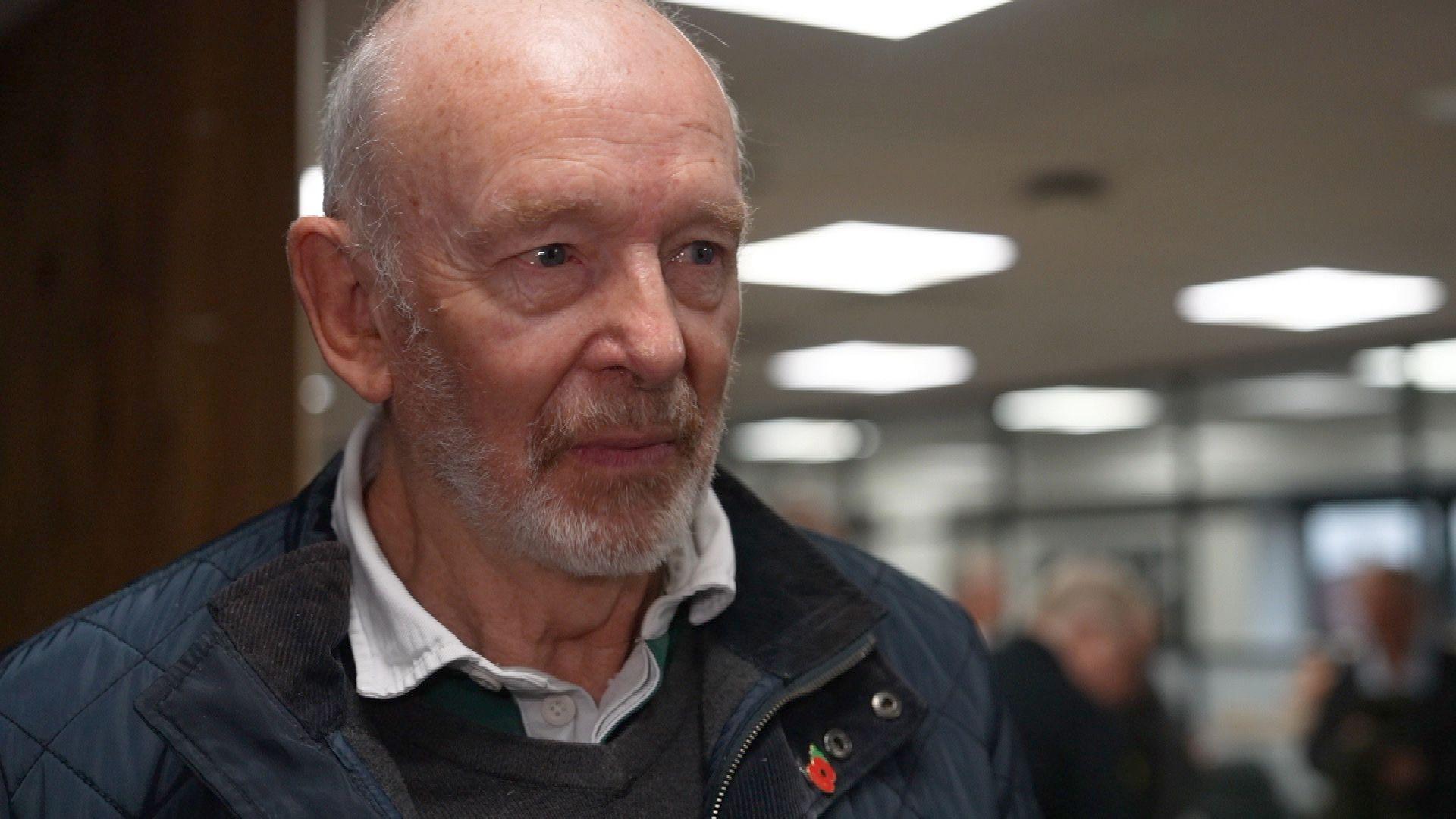 A head and shoulders picture of a man looking off-camera staring at the camera, wearing a white shirt with a blue coat over it. He has a grey beard and is wearing a metal poppy badge on his lapel. He is indoors, but the room behind him is blurred.