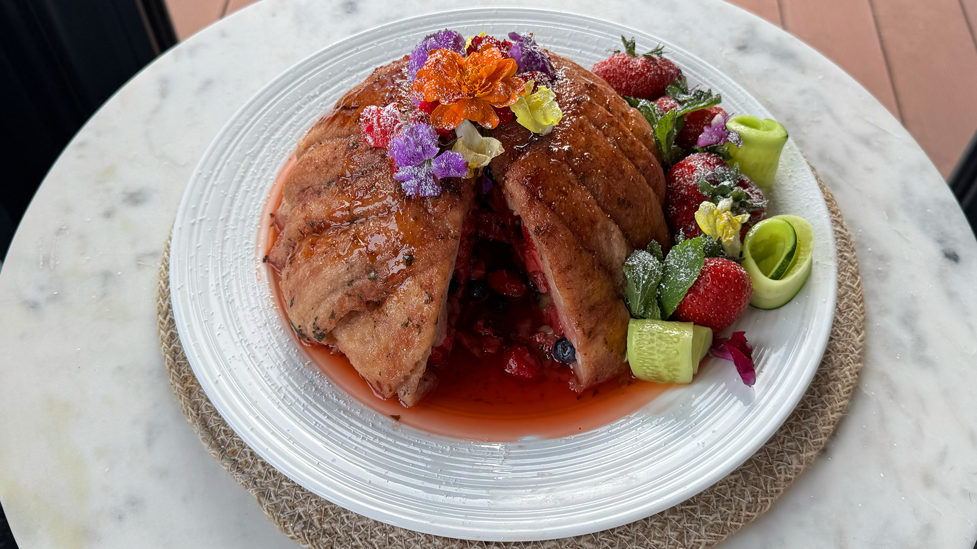 Yvonne's Wimbledon summer pudding. A dome of sweet juice-soaked bread, sliced open and revealing the red and blue berries inside. Garnished with purple, orange and yellow flowers on top, with juicy red strawberries and green twirls of cucumber and fresh mint around the side. All sitting on a white ceramic plate.