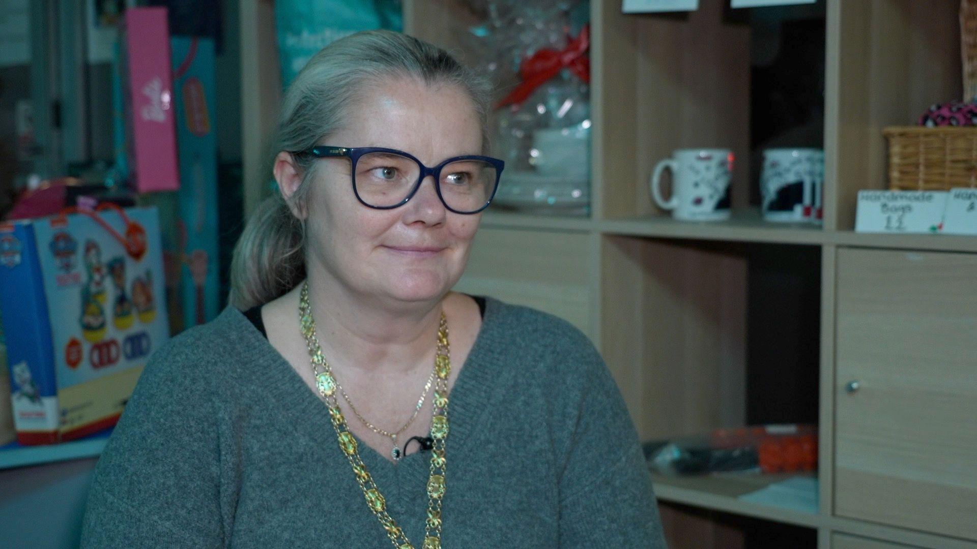 Emma Gilman, who has dark glasses, is photographed wearing a green v-necked jumper, and a gold-colour chain.