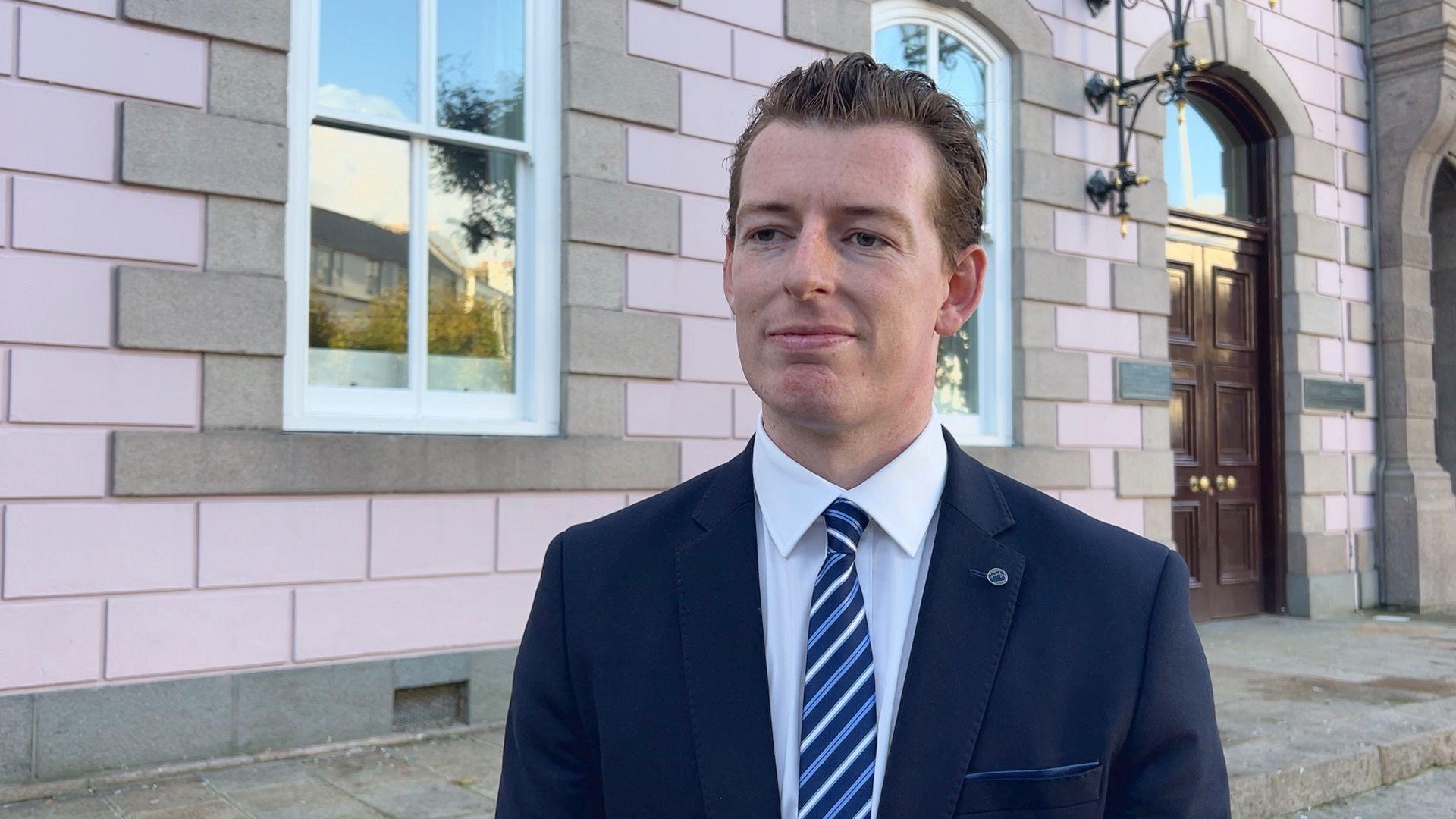 Deputy Max Andrews standing outside the States Assembly building, which as light pink walls and large windows. He is wearing a dark blue suit with a white dress shirt and a blue-and-white striped tie. He has short brown hair, neatly styled, and is looking slightly to the side with a composed expression. 