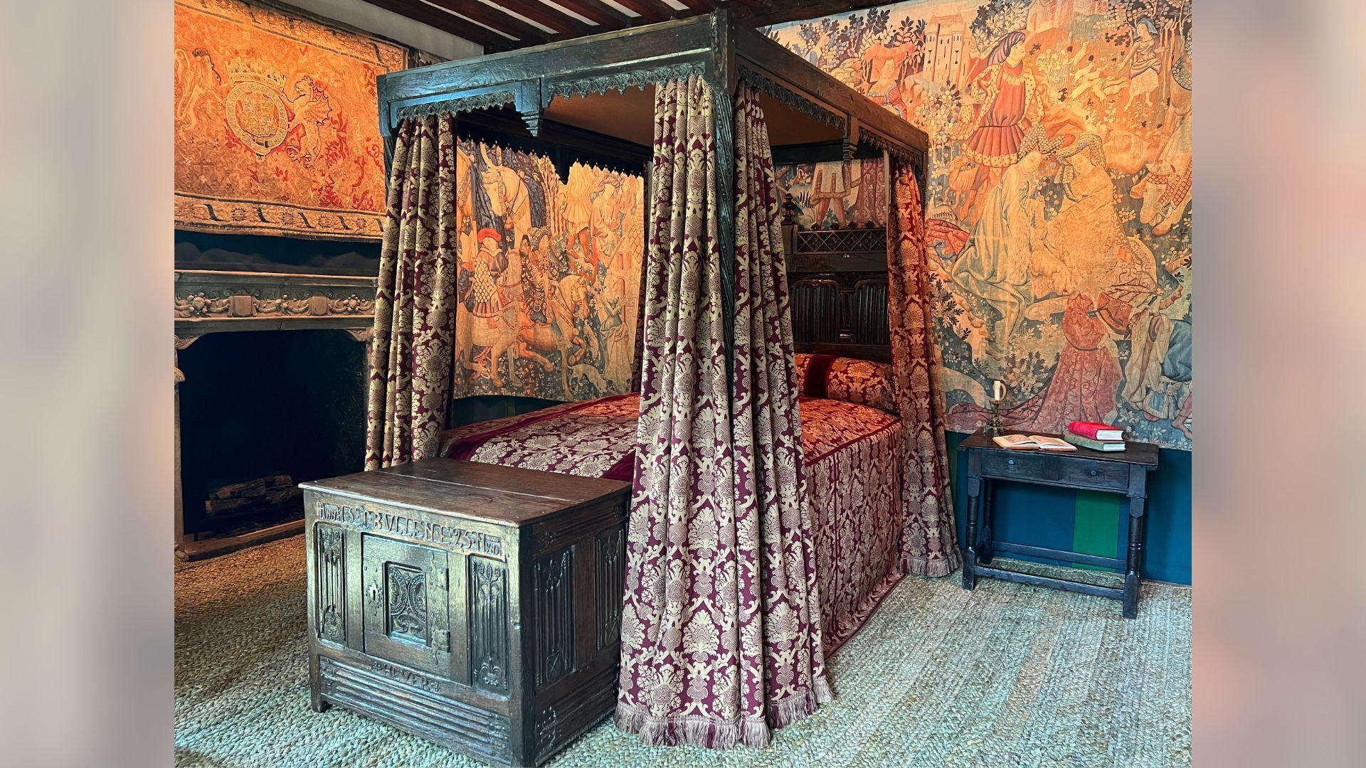 An orange room with a four-poster dark wood bed. There are red and gold curtains hanging from the corners. There is a dark wood chest at the foot of the bed. The carpet is light green.