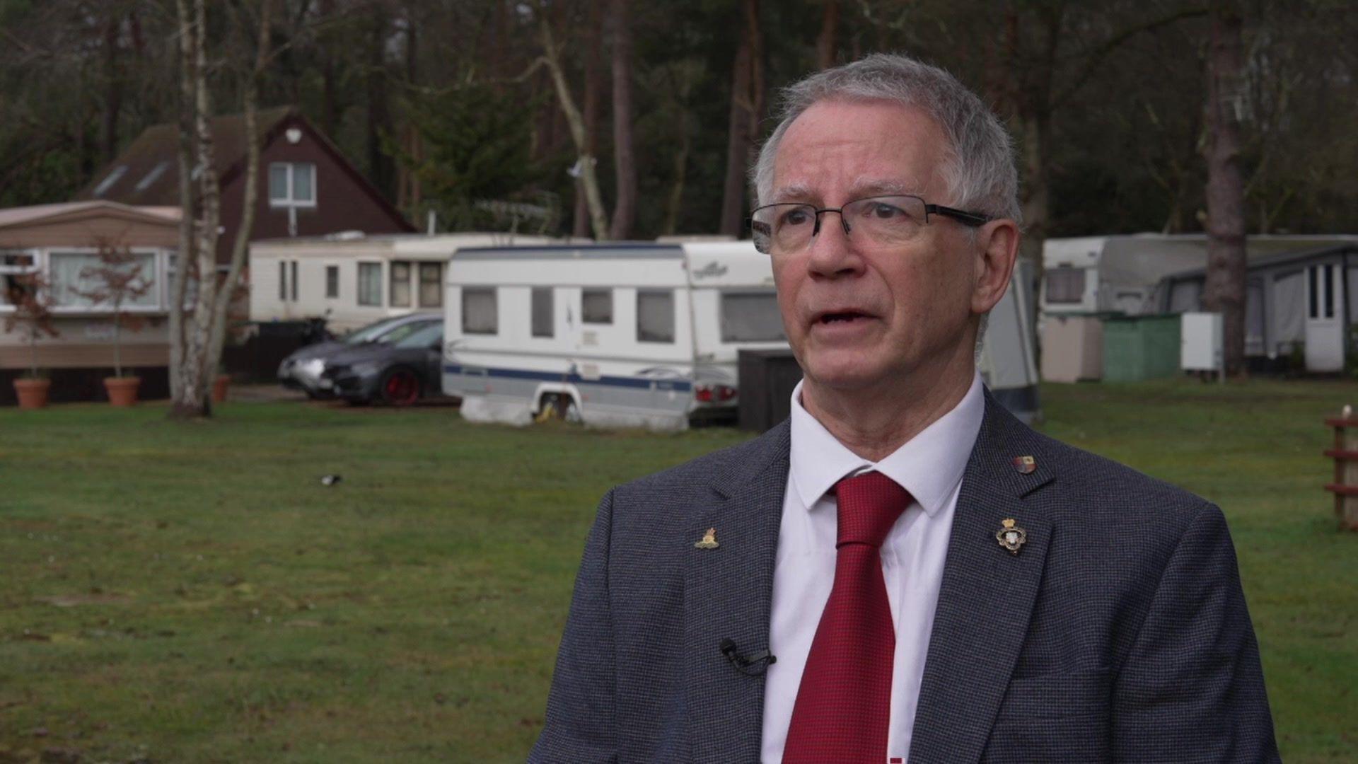 A man with grey hair and glasses, wearing a suit with a red tie. He's looking straight at the camera and there's a row of caravans behind him.