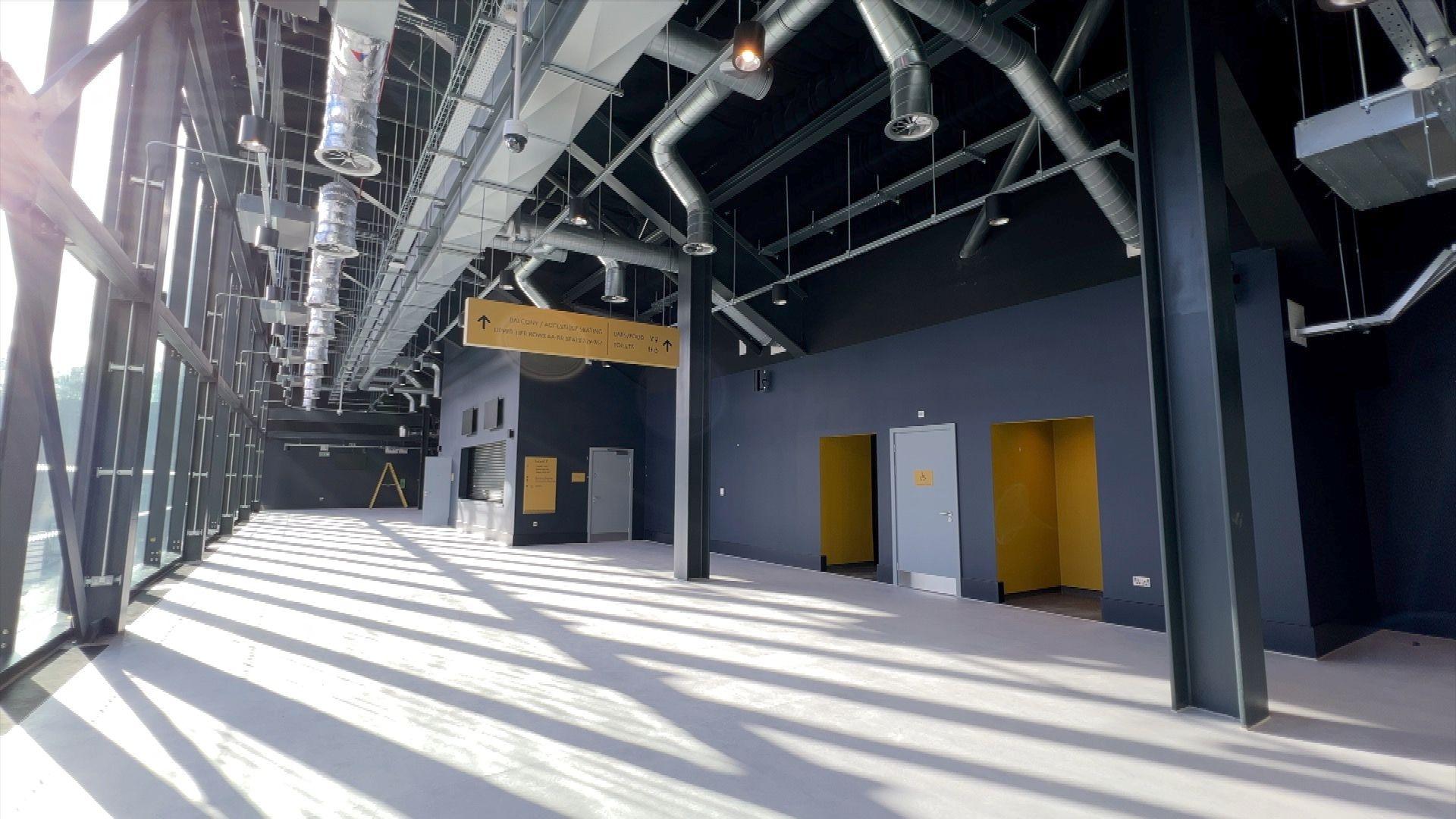 A concourse inside of the arena, with glass windows on the left and doors into the arena on the right. The walls and doors are grey or yellow  