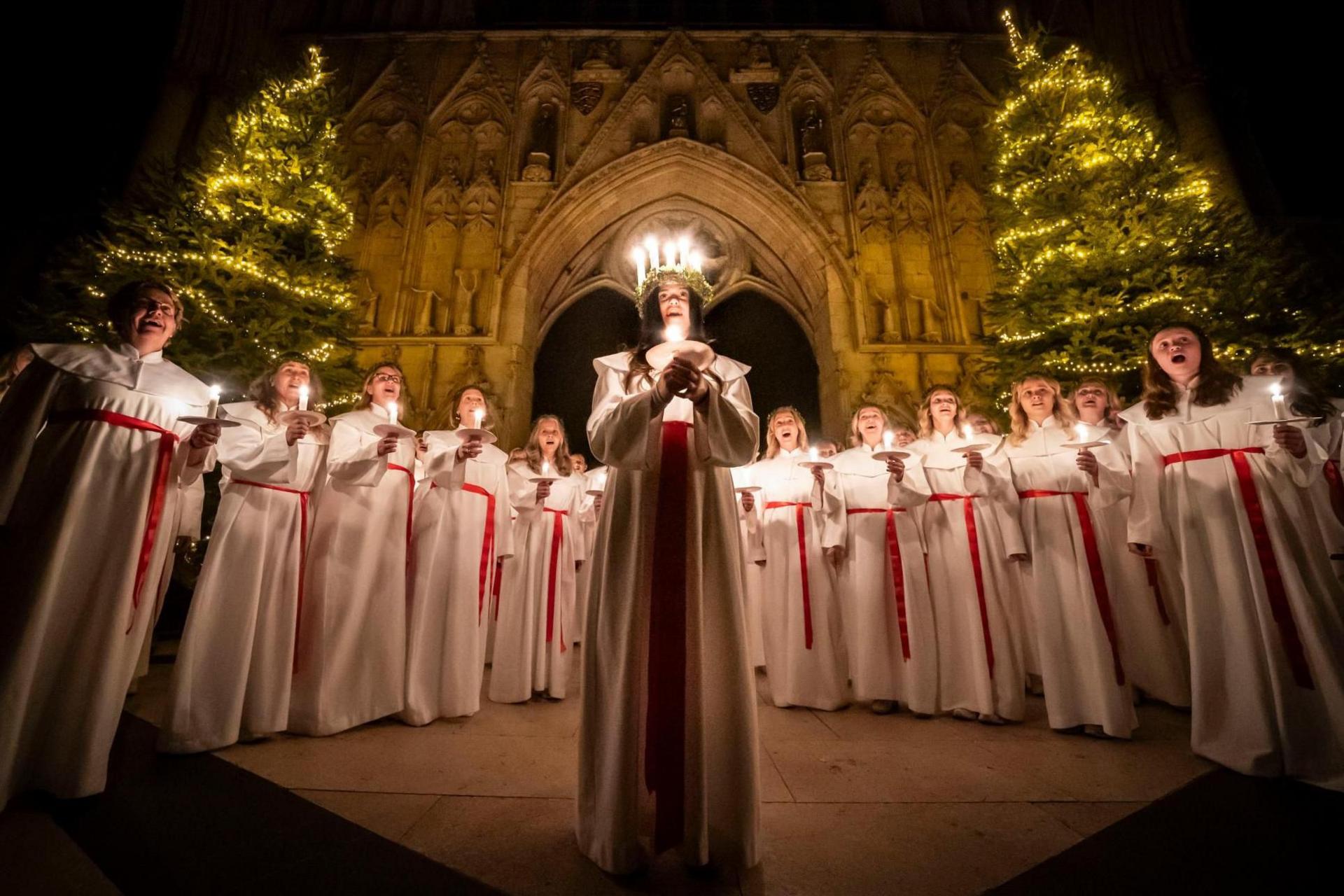 A young woman wearing a crown of lighted candles and holding another candle with both hands, sings surrounded by other members of the female choir, who are also holding candles. Behind them at York Minster are two Christmas trees, decorated with lights.