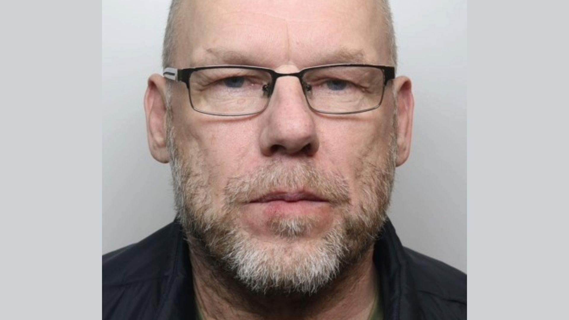 Police mugshot of Dean Hearson. He is a middle-aged looking man, with thinning hair, wearing glasses and has a light coloured moustache and beard. He is wearing a dark shirt.