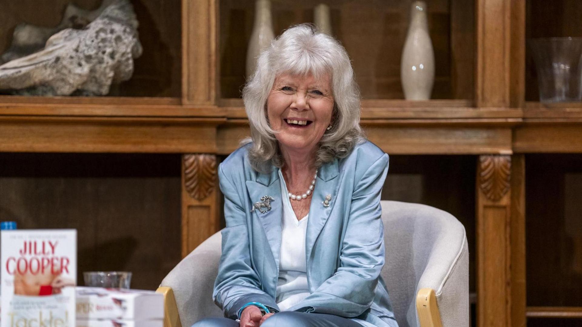 Jilly Cooper, wearing a light blue jacket, sits on a beige chair at a book event.