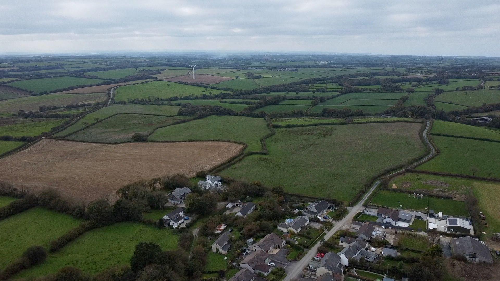 An ariel view of gently rolling green fields and countryside. Part of a hamlet is visable in the foreground, with fields into the distance. One of the fields has a wind turbine.