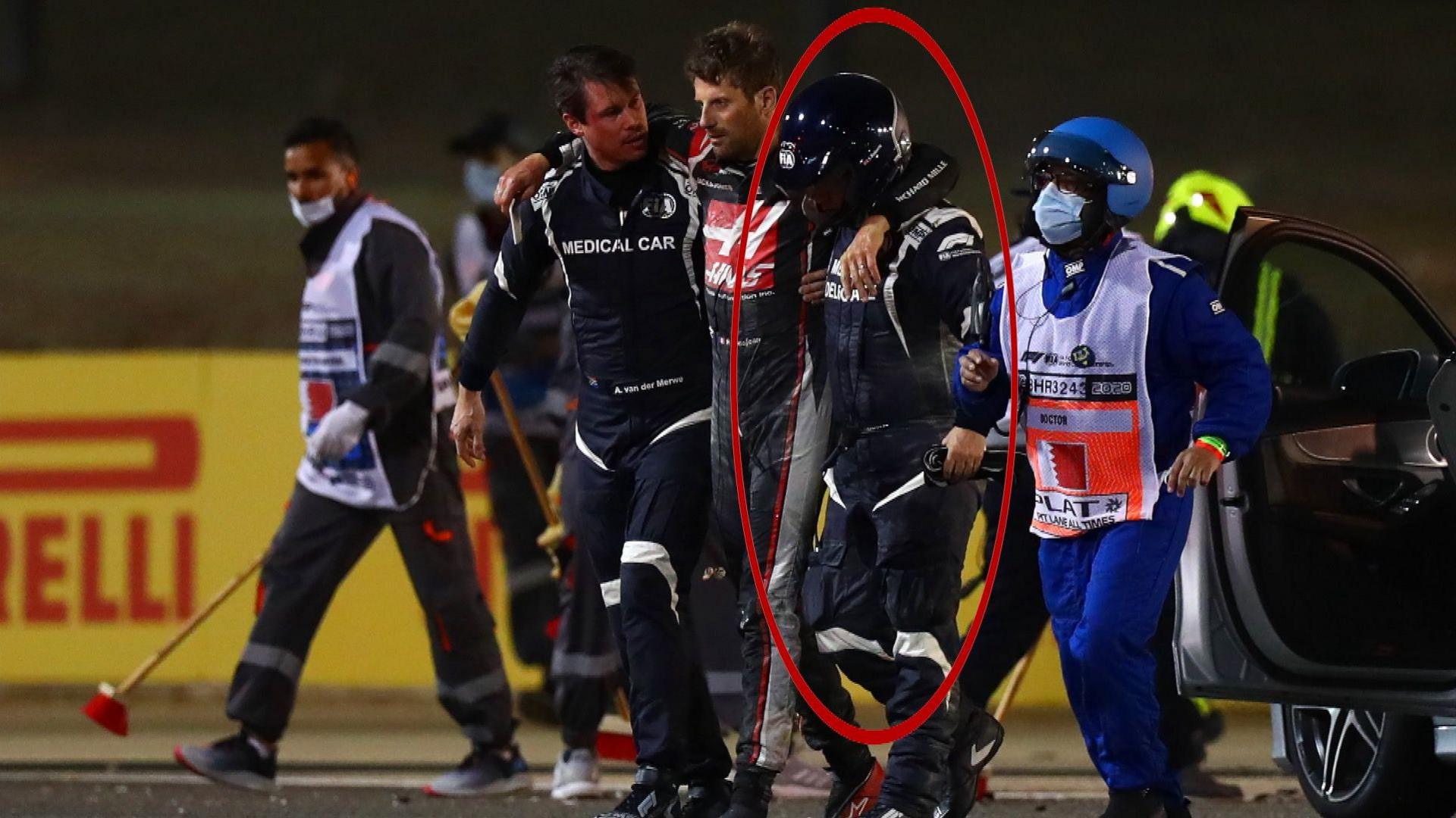 A man with dark hair wearing a blue jumpsuit is supported by two other people to walk to the left of the screen while other people can be seen around them.