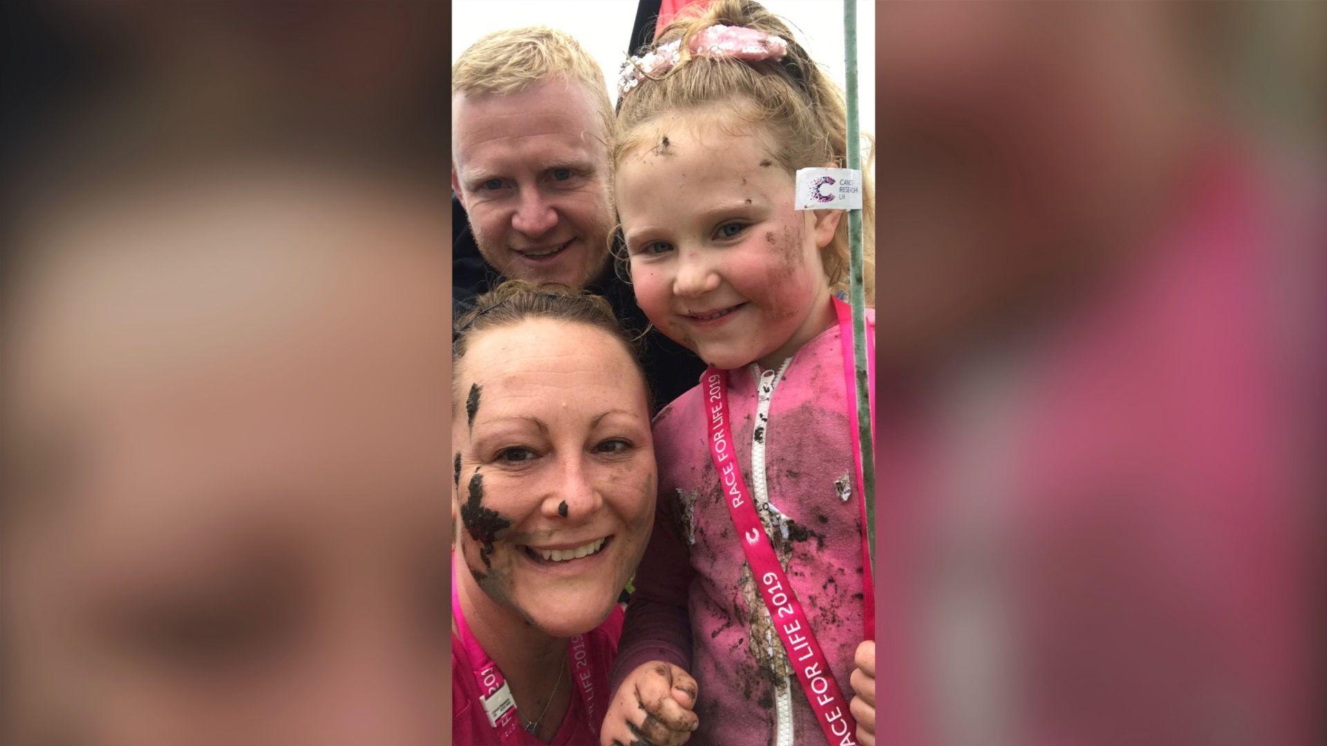 Lee, Michelle and Connie pose for a photograph, smiling into the camera. They have mud on their faces and are wearing pink Race for Life 2019 lanyards around their necks.