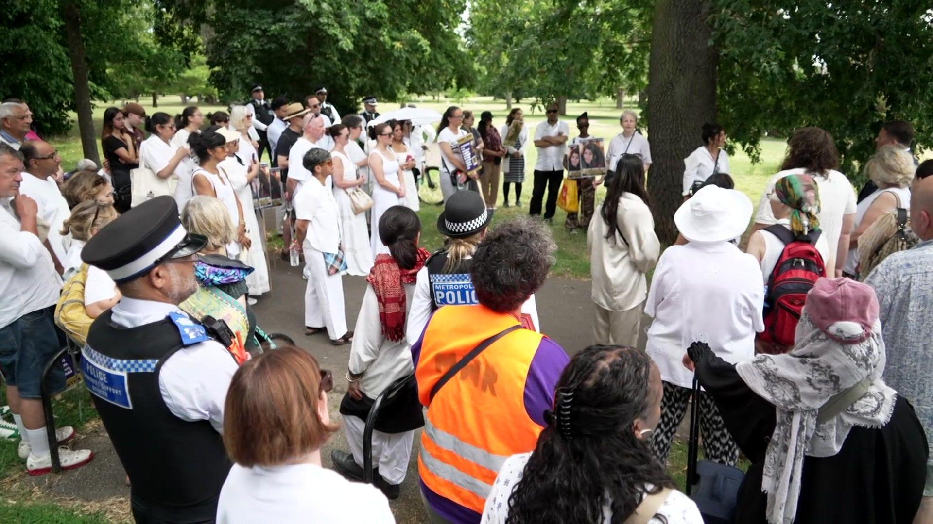 A large group gathers in a park with police officers present, listening during a memorial event for Zara Aleena.