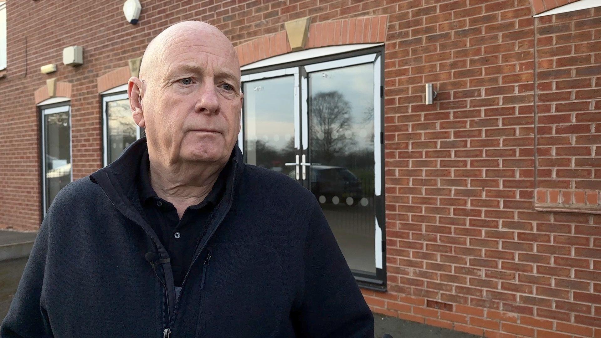 Head and shoulders shot of Steve Lowbridge with a brick house and patio doors in the background. Steve is wearing a dark fleece and dark polo shirt underneath
