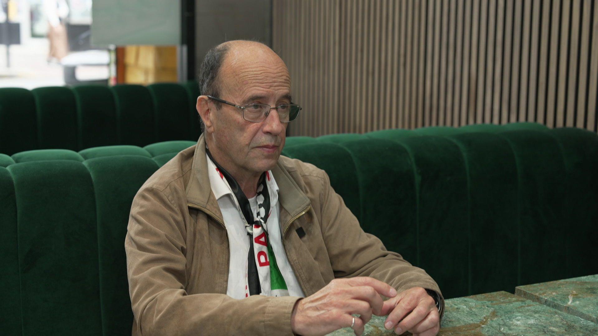 Jonathan Fluxman in a white shirt, Palestine flag scarf, and brown jacket.
