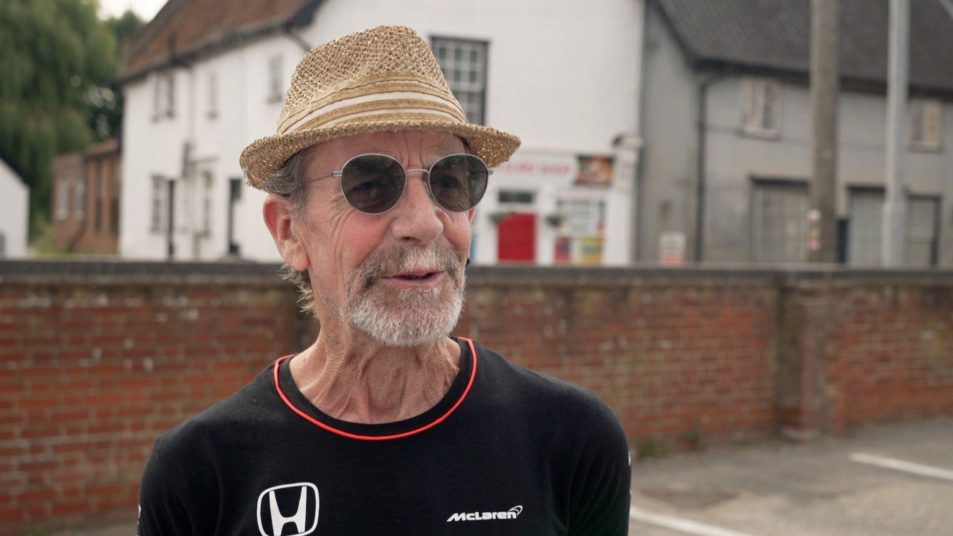 A man with a goatee beard wearing a black T-shirt, a straw pork pie hat and sunglasses stands in a car park with a wall and a shop behind him.