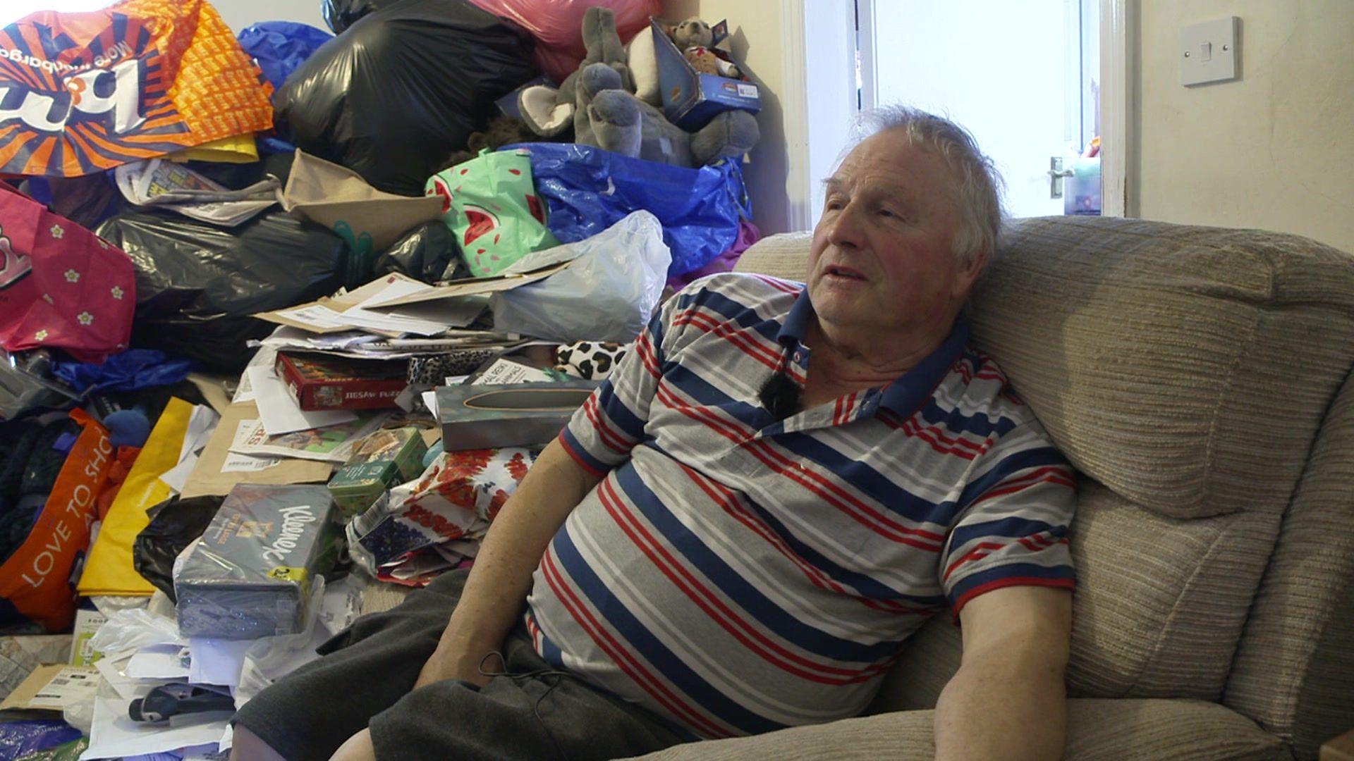 John Wilson sitting in his front room surrounded by clutter