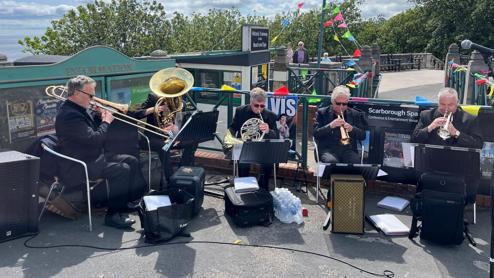 Five people sitting in chairs outside playing musical instruments. They are looking at sheets of music on stands with speakers in front of them. In the background there are railings which are decorated with colourful bunting.