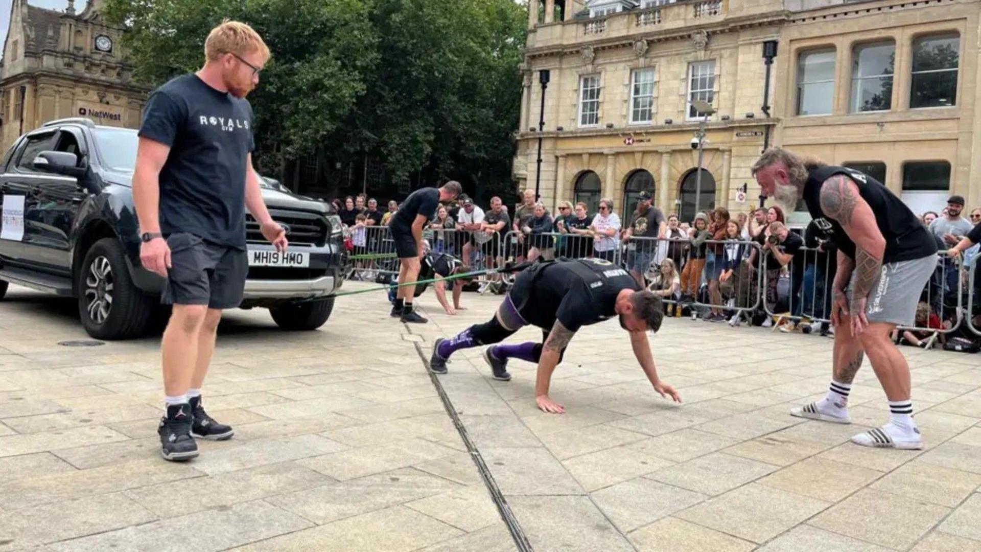 Two men on their hands and knees with bands attached to their waists are trying to pull trucks along the Cathedral Square. Onlookers behind metal barriers are watching them while three event organisers are observing and encouraging them.