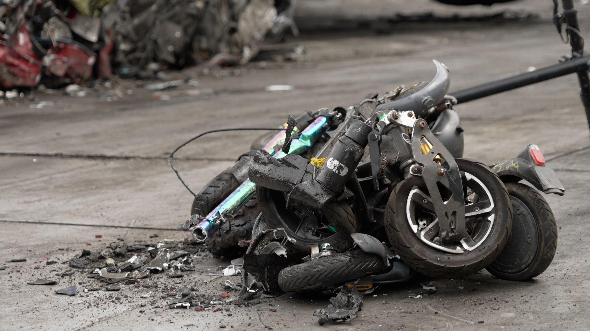 The image shows a pile of crumpled up e-scooters and e-bikes. They are all black in colour apart from one which is a two toned green and purple colour. 