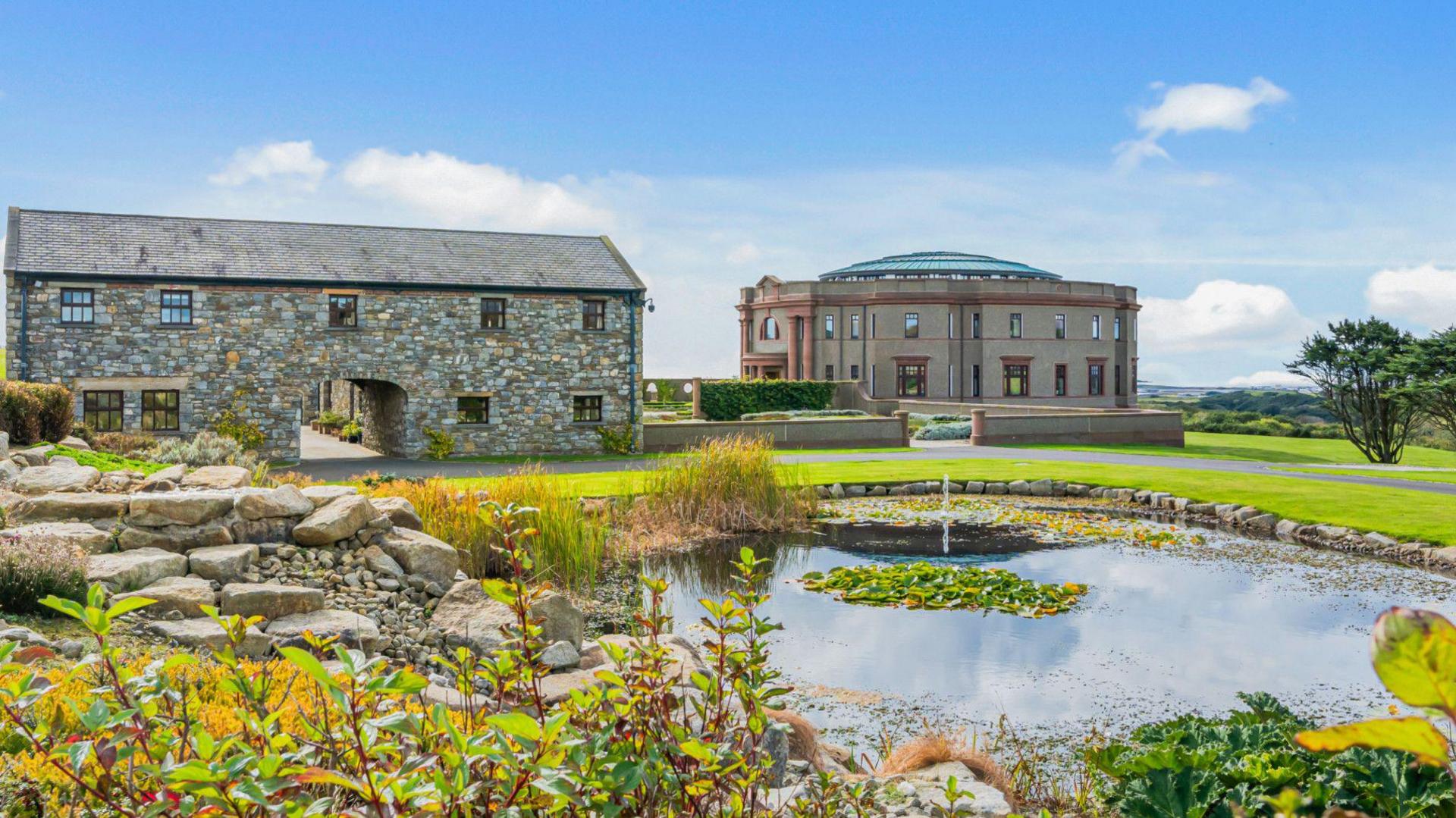 The property with a large farm style building to the left and a large pond with a water feature in front.