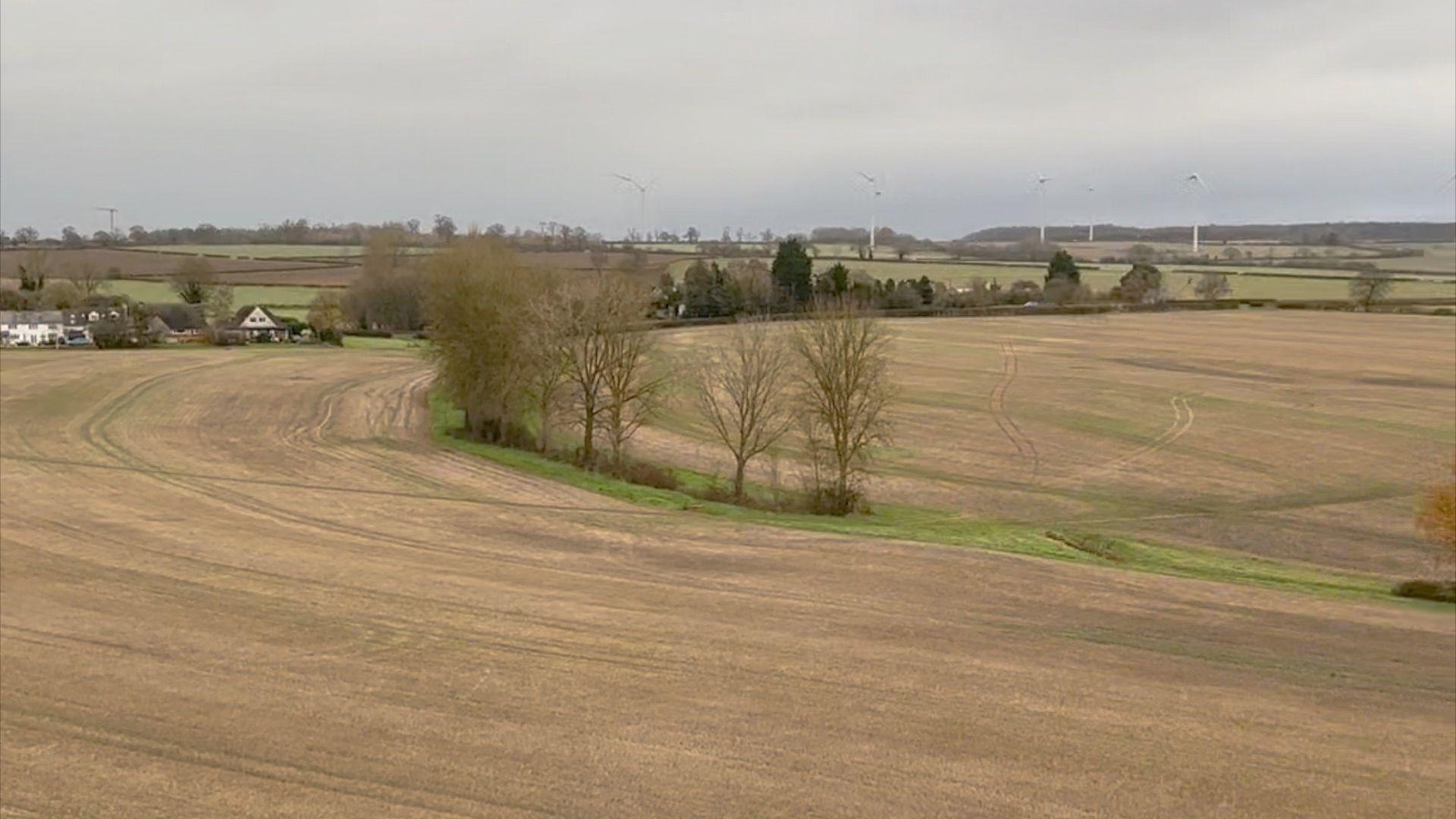 A wide shot of the view from the tower, showing brown fields, trees and general countryside. There are some wind turbines in the distance.