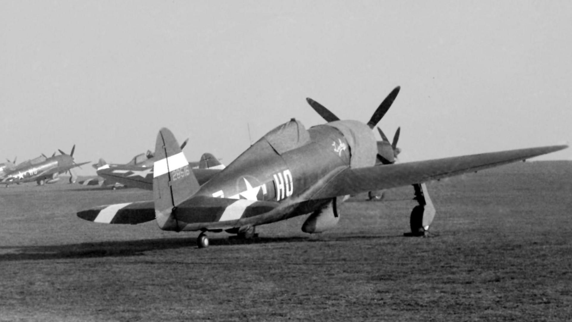 A black and white photo shows an old aeroplane, with its propeller facing away from the camera. It is seen parked up on a field, with other similar planes in the background.