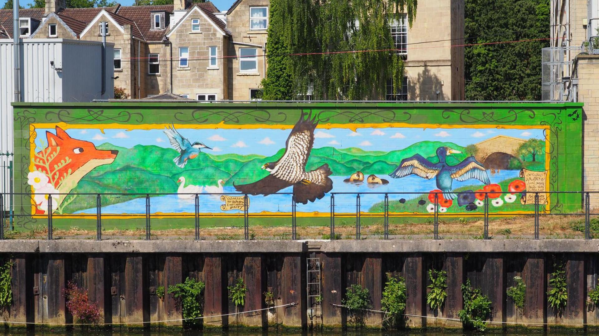 Colourful mural of animals and a river, painted on the side of a building, with houses behind it. The mural shows a fox, a duck, swans, fish, a kingfisher, otters and a bird of prey. It includes a river with a bridge over it that joins two sides of grassland.