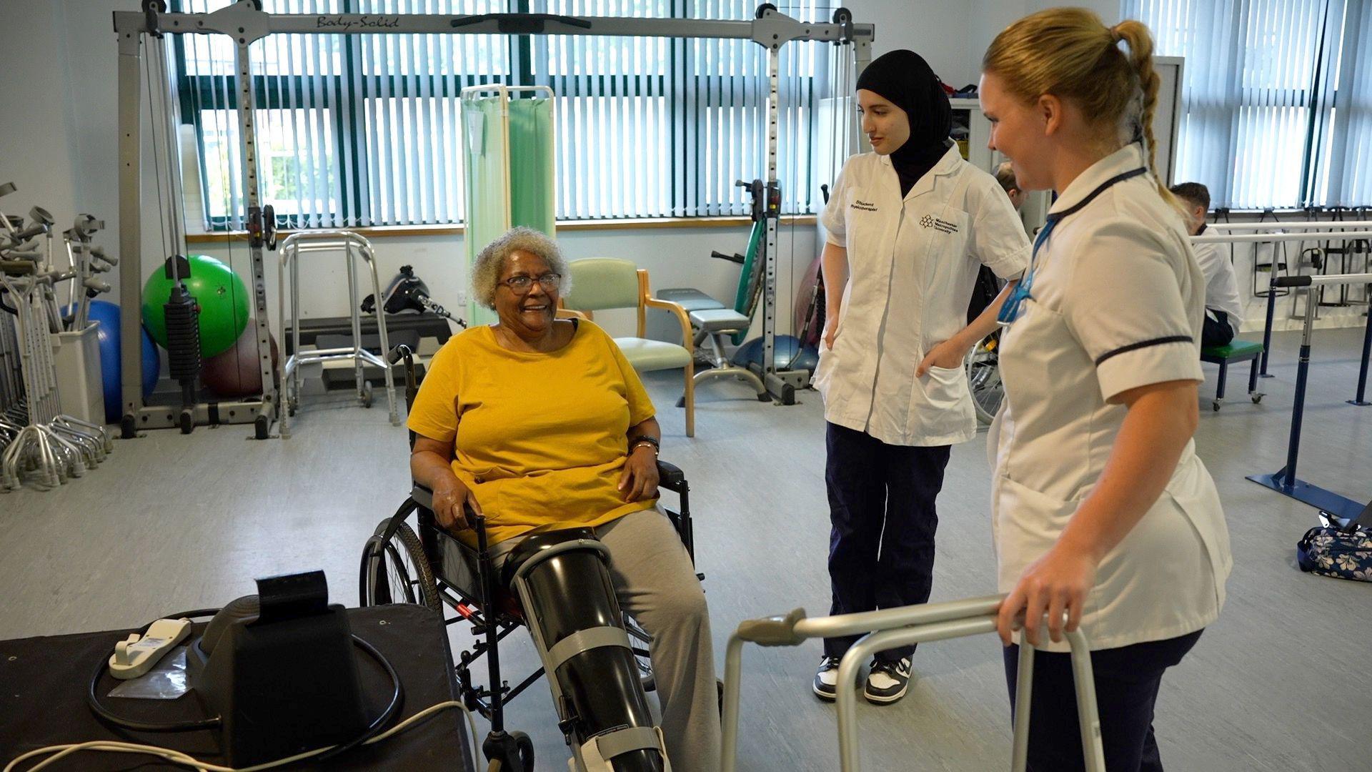 A woman wearing a yellow t-shirt is sat in a wheelchair and smiles up at two nursing staff.
