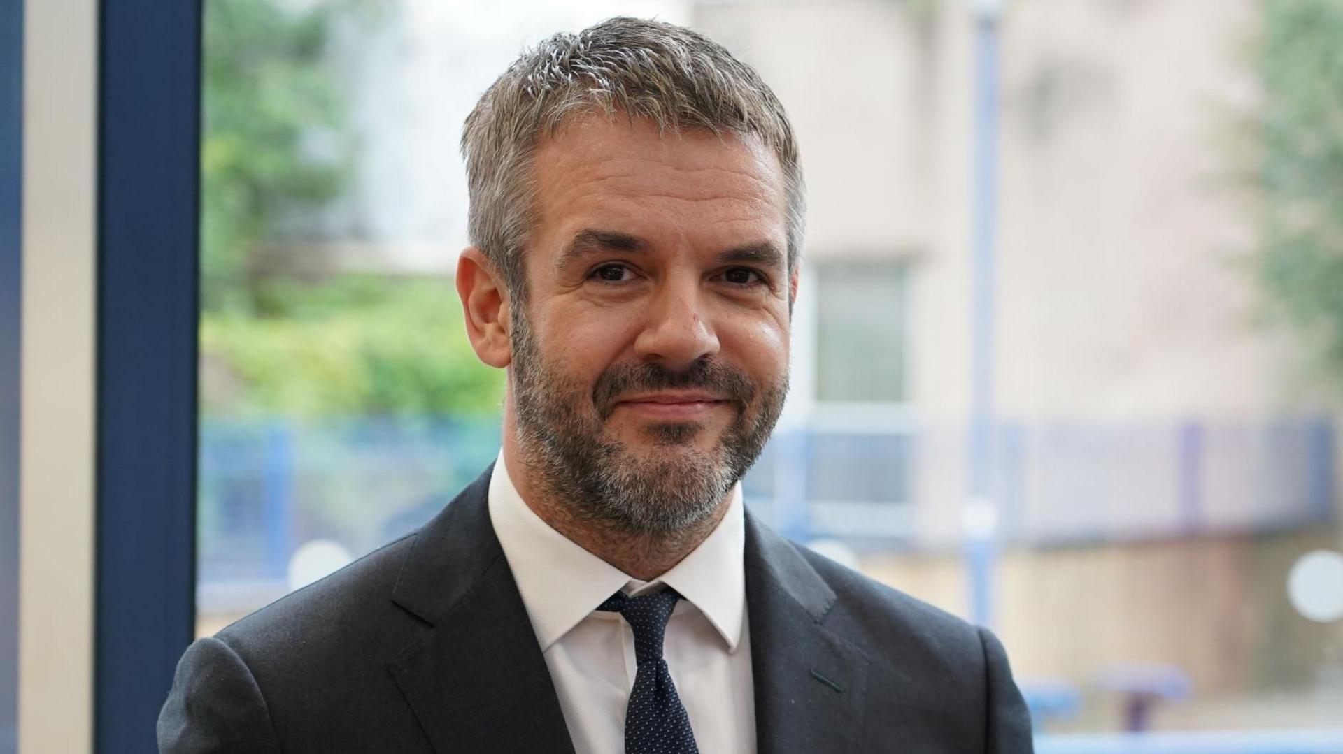 Coppard wears a black suit and tie as he smiles at the camera. He has salt and pepper hair and facial stubble.