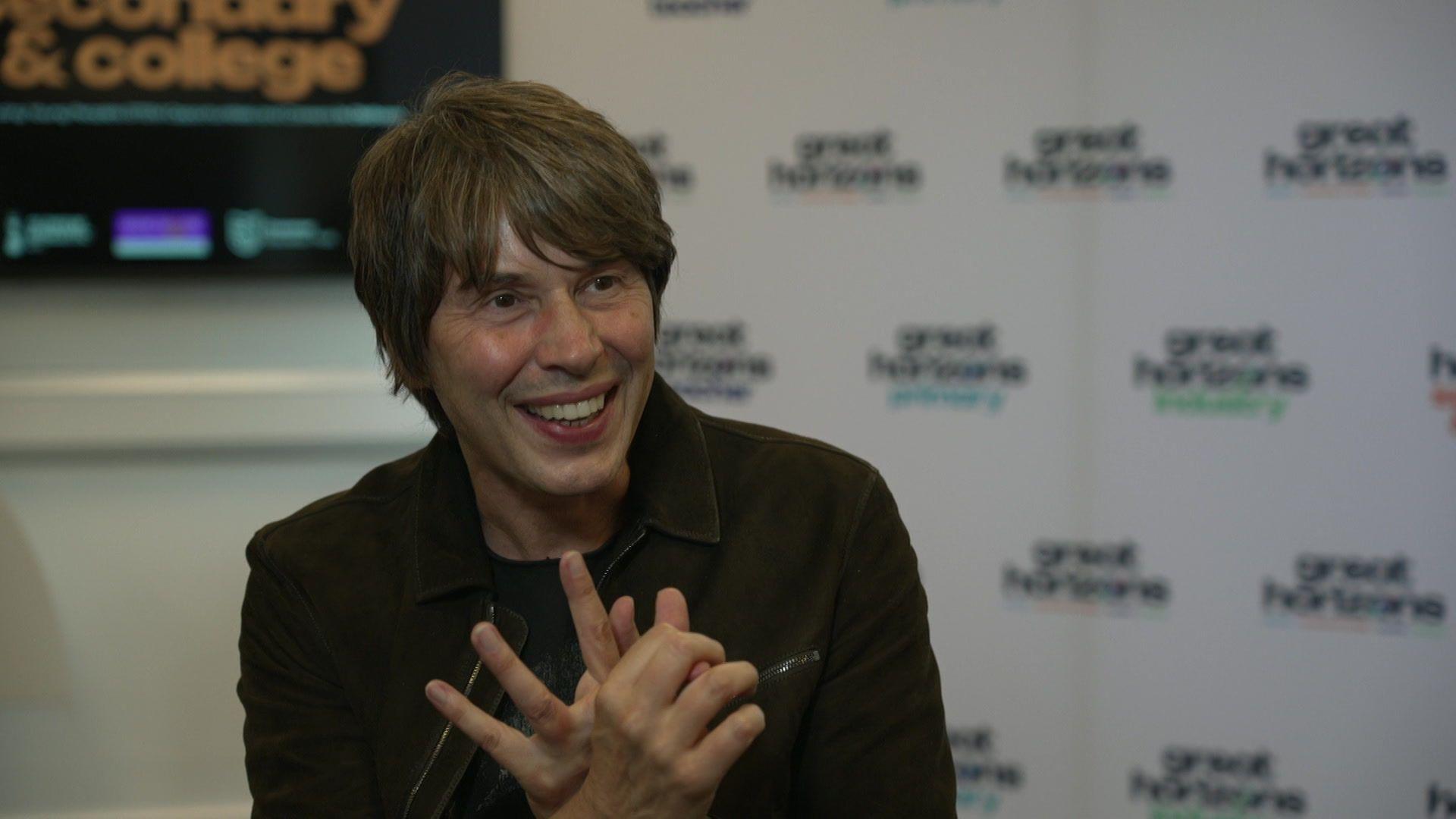 Professor Brian Cox sits in front of a white background. He wears a black jacket and a black T-shirt. He has collar-length dark hair and a sweeping fringe.