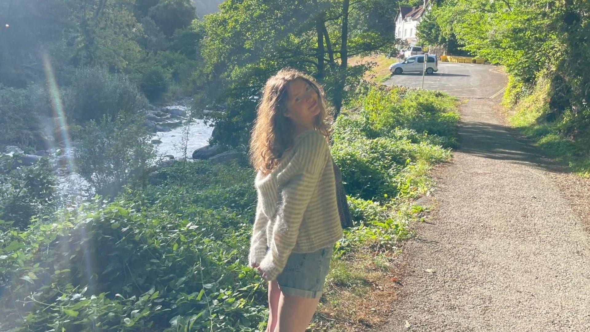 Emily wears denim shorts and a jumper, and is standing on a path beside a stream, bordered by greenery, while the sun shines in the background