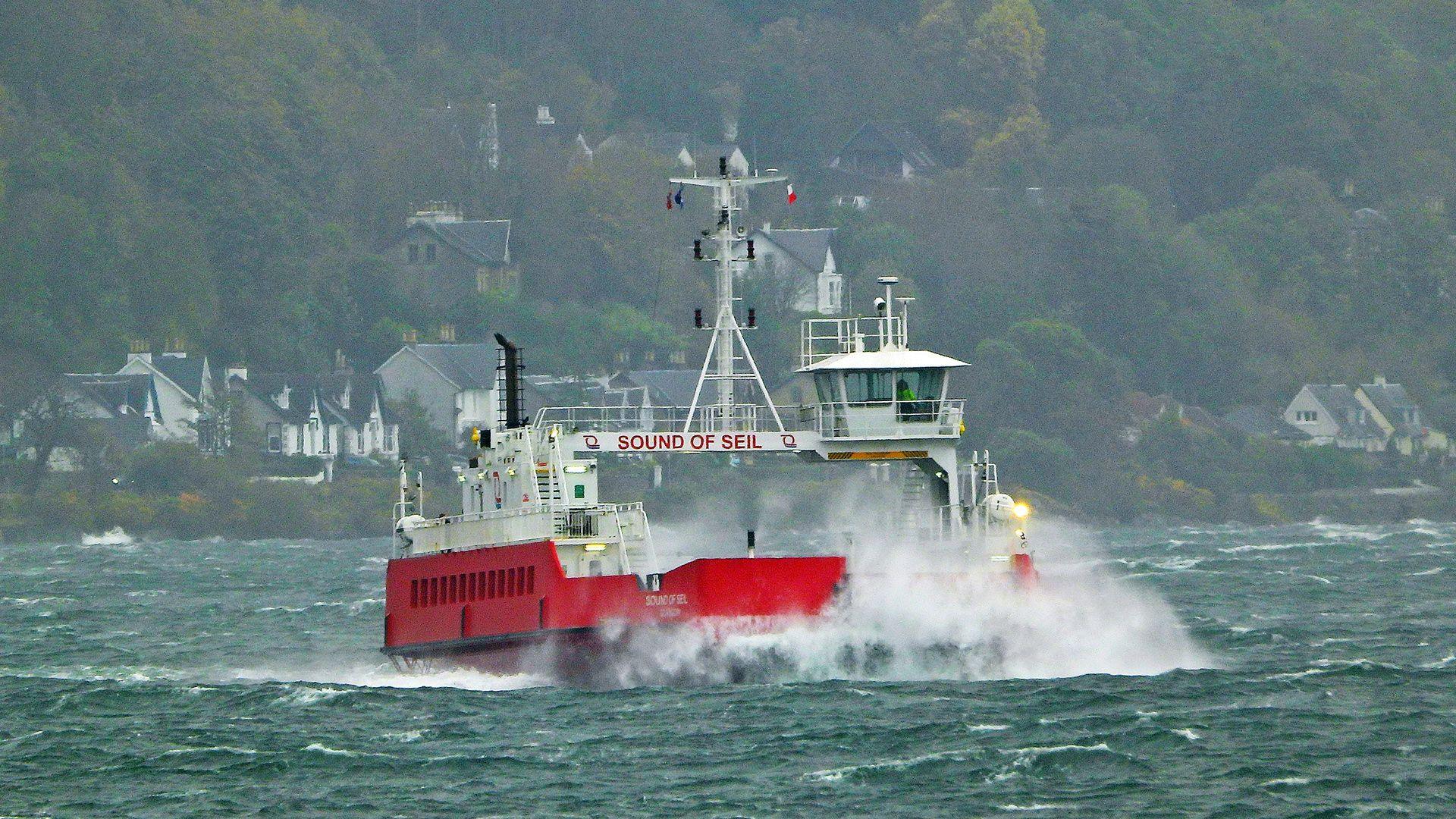 large waves hit bow of car ferry at sea, with houses and trees on shore in distance