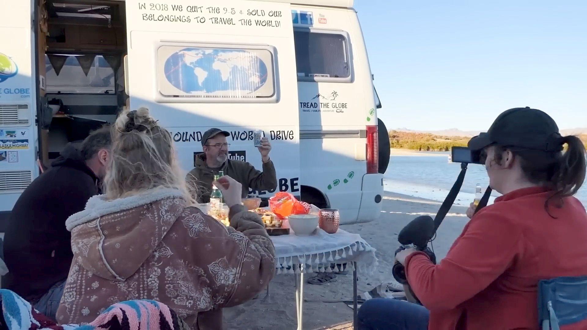 A group of four people sit at a small table in front of a white camper van, there are bowls on the table and some food items