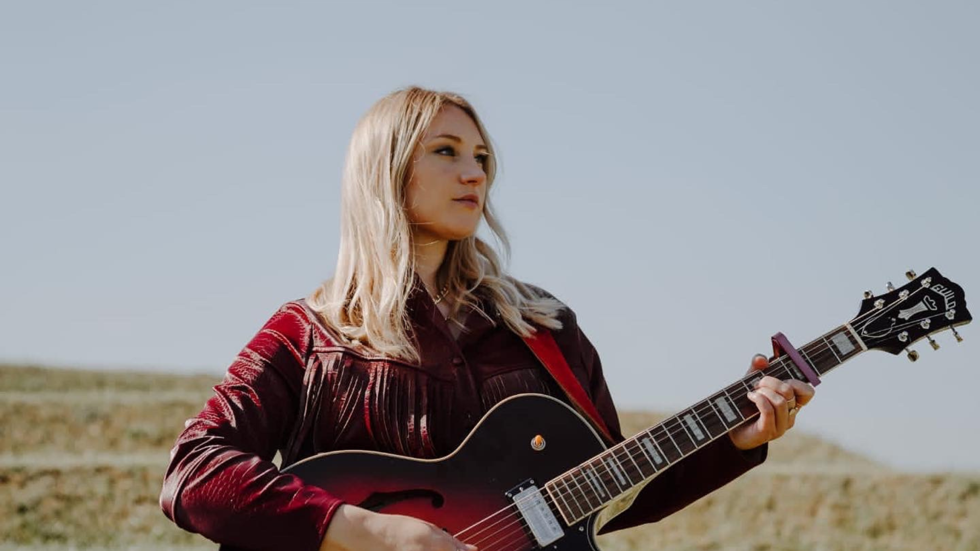 Dolly Mavies in a dark red jacket standing in a field holding a guitar