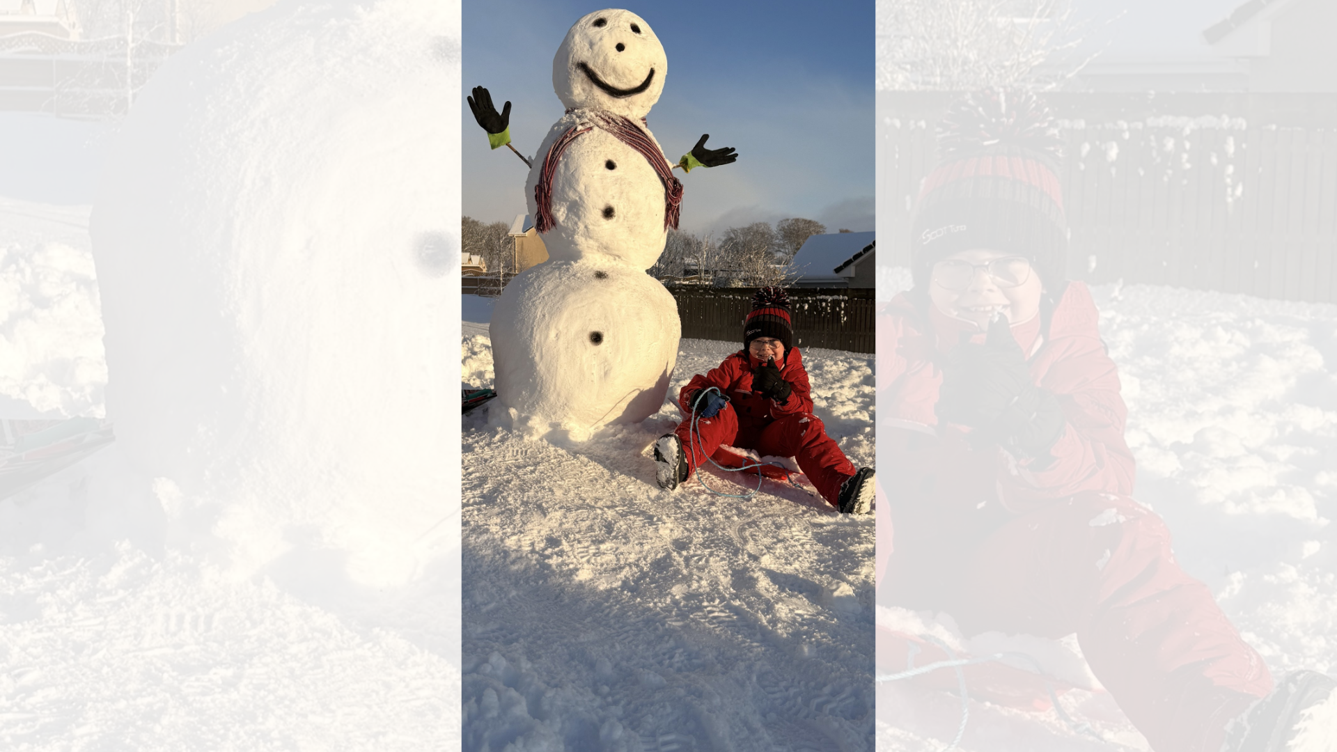 a boy in a red jumpsuit and black and red beanie sitting in the snow next to a big snowman with a smiley face.