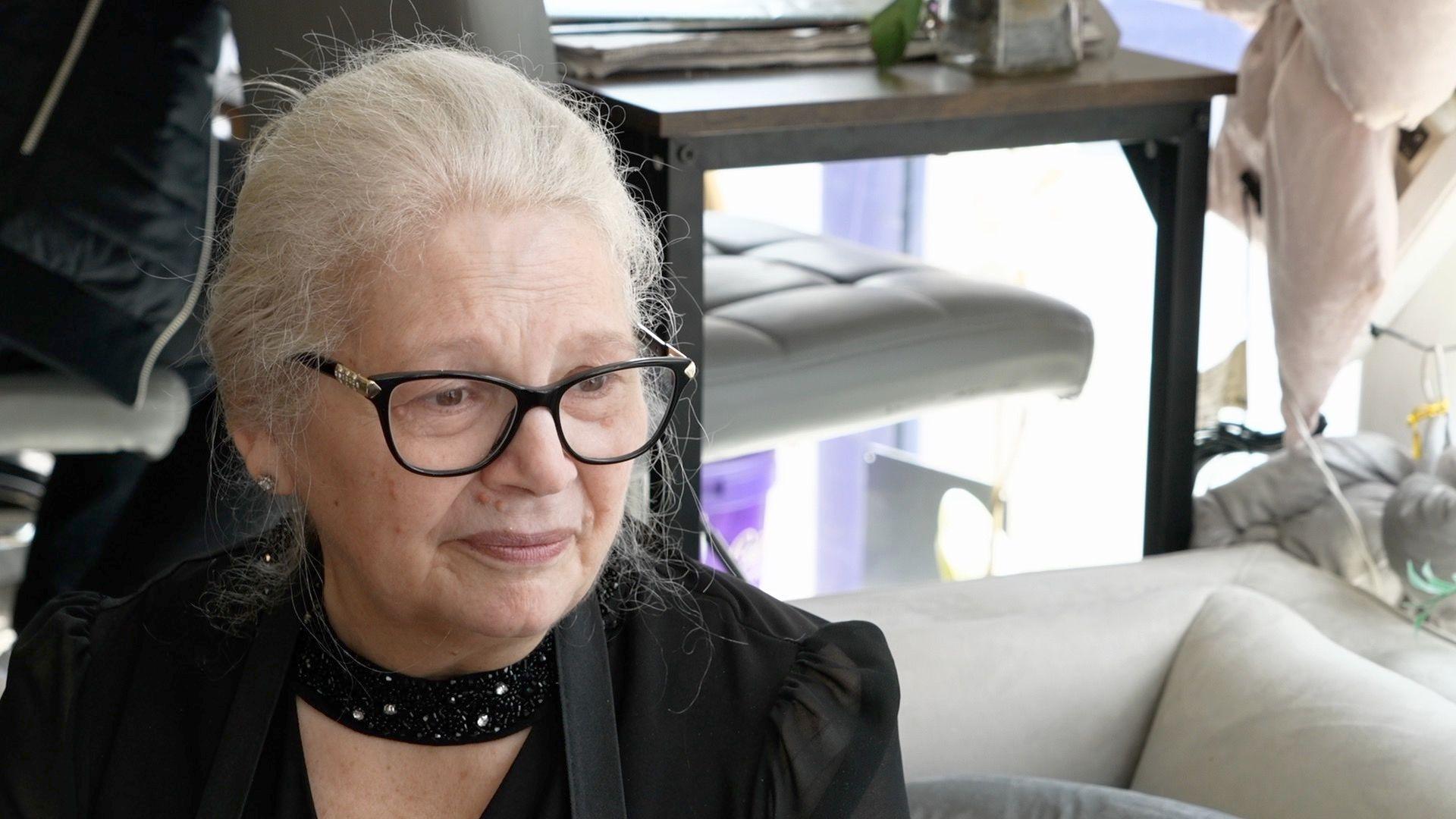 Francis Newson smiles as she looks away from the camera while sitting on a white sofa. She has grey hair that is tied back, glasses and wears a black scarf and black blouse.